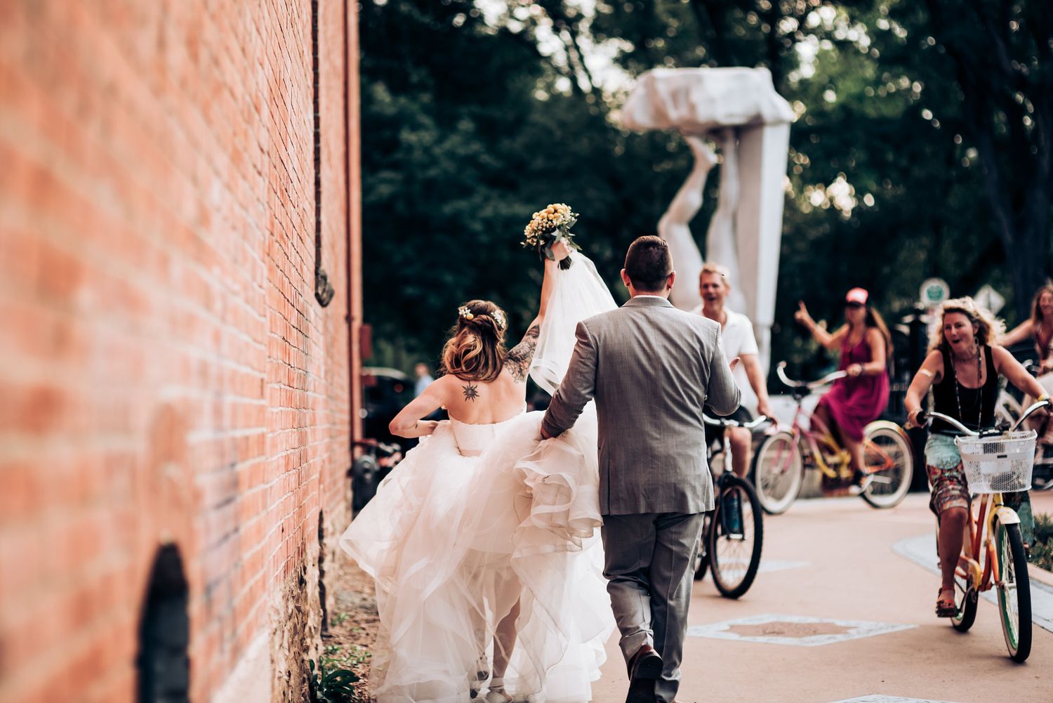 Candid moment of wedding couple walking past brick wall as cyclists pass by.