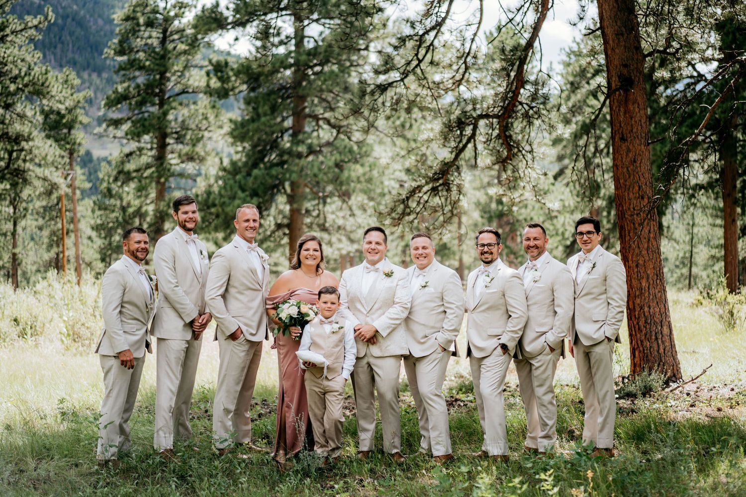 Wedding party in light colored suits poses together in a mountain forest setting.
