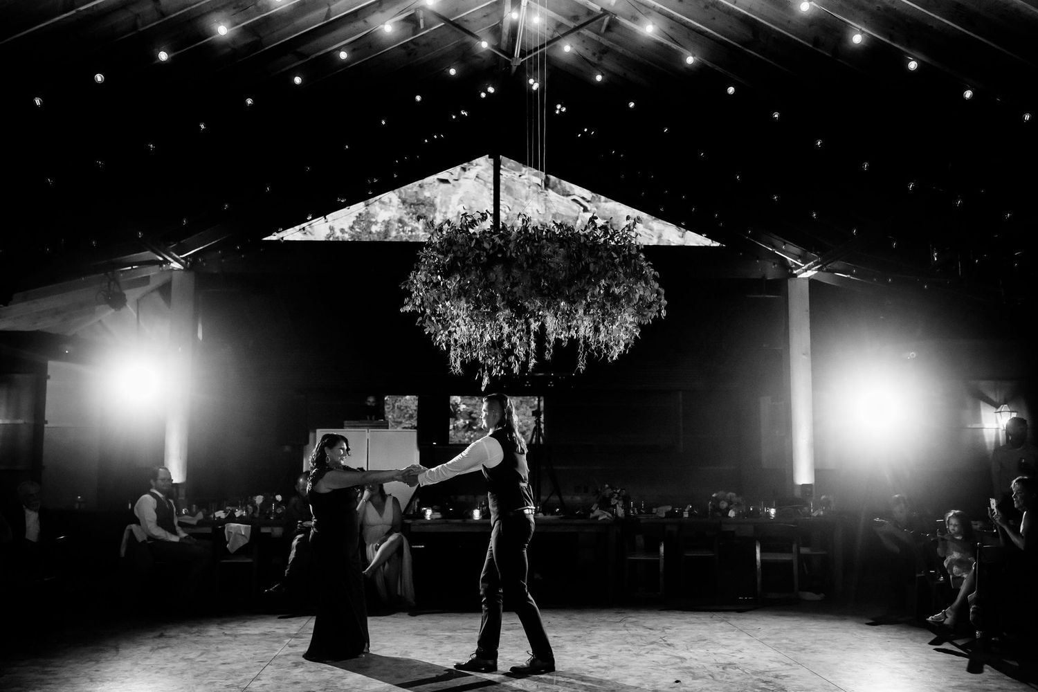 A black and white photo of a couple's first dance under a rustic pavilion with string lights.