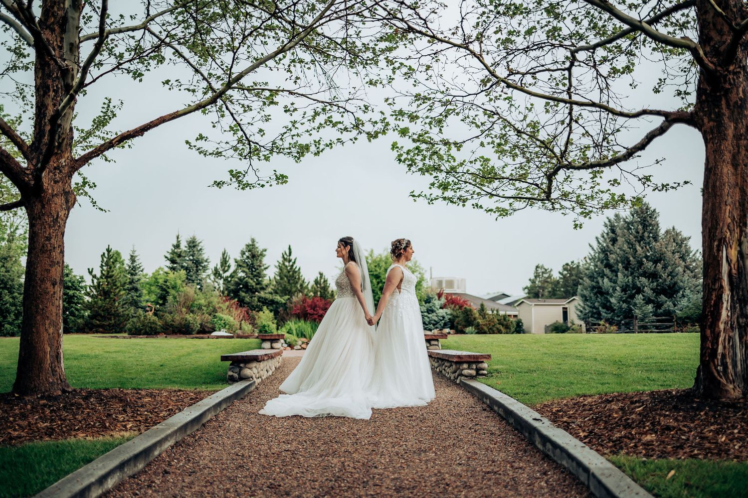 Two brides in white dresses stand together on a tree-lined garden path.