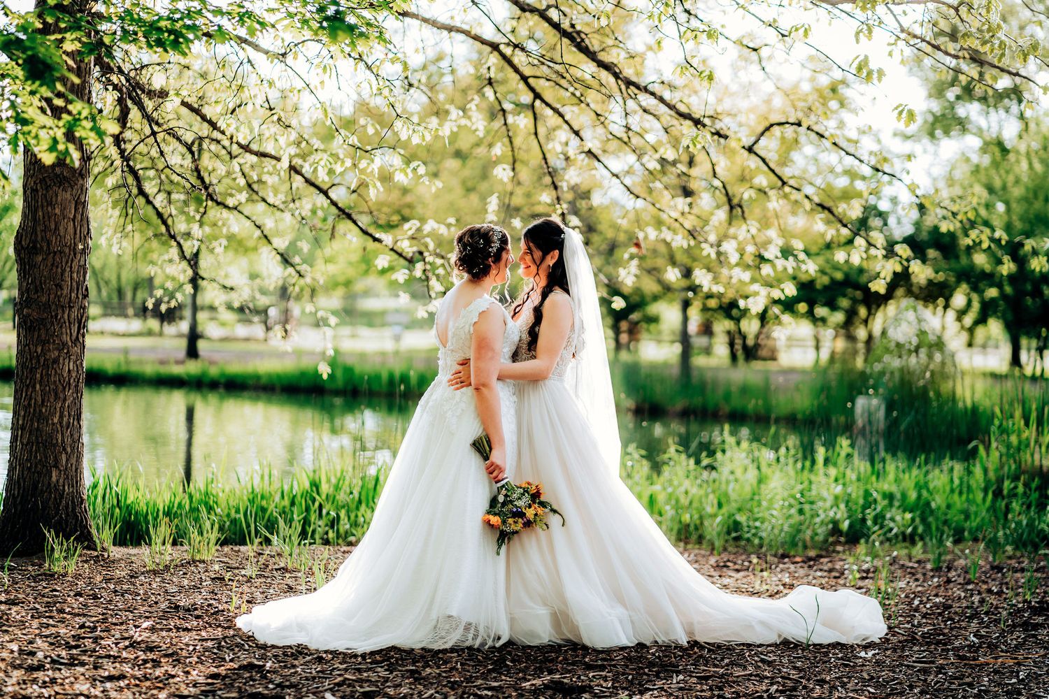 Couple embracing under blooming trees near a pond in wedding attire.