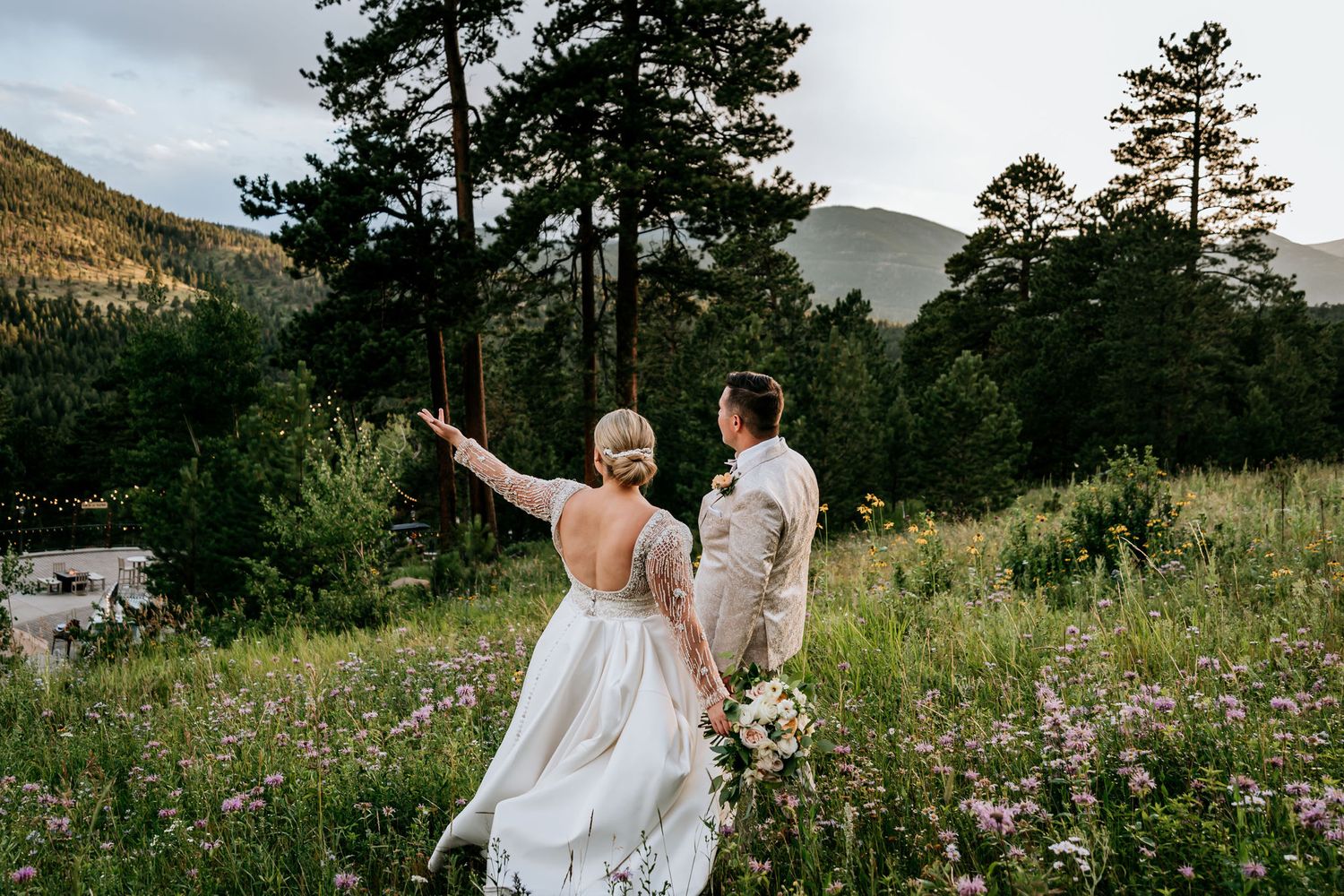 A couple celebrates with arms raised in a mountain meadow filled with wildflowers at sunset.