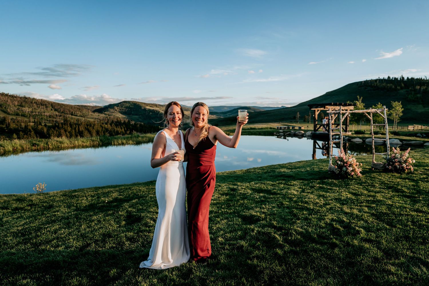Two people pose against scenic mountain backdrop with lake and wooden arch at outdoor venue.