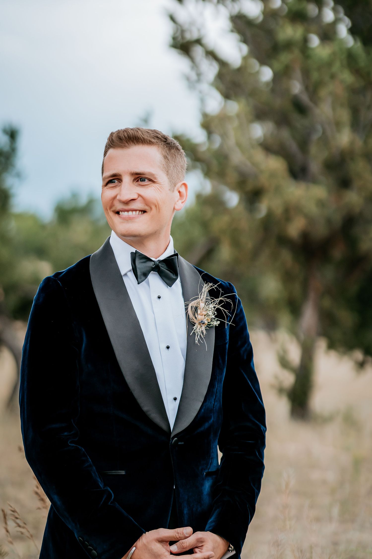 A groom in a classic black velvet tuxedo smiles during an outdoor wedding.