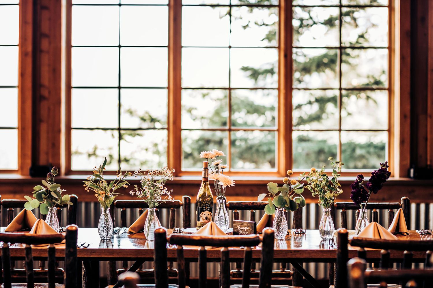 Light-filled reception venue with wooden tables and chairs set for a wedding celebration.