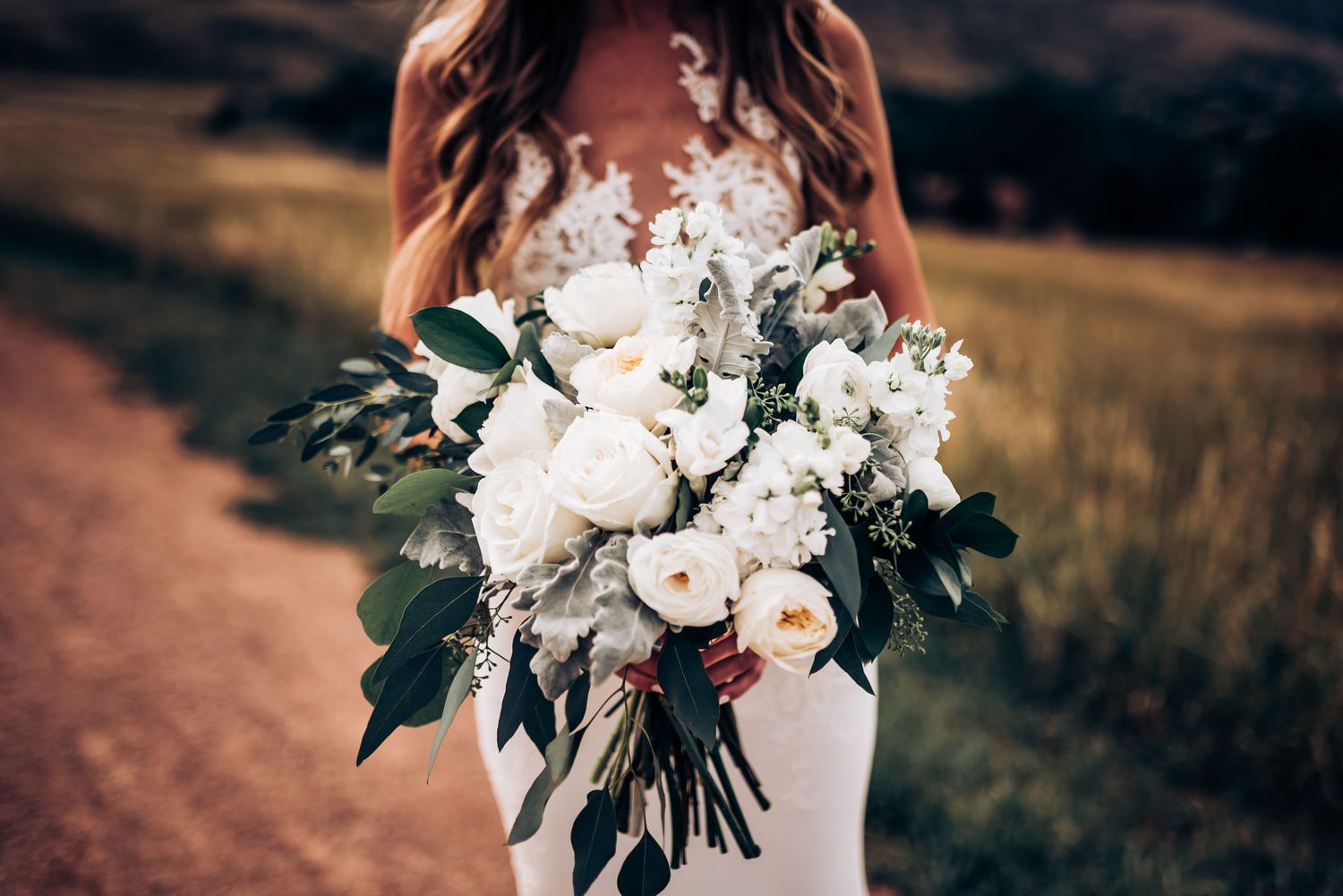 Close-up of an elegant white bridal bouquet with green foliage held against a white wedding dress on a dirt path.