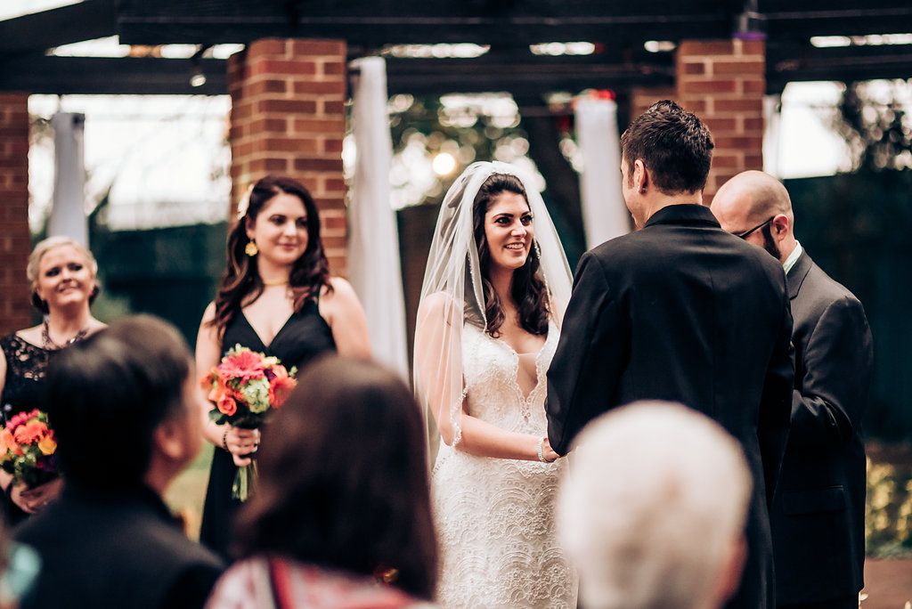 Wedding ceremony outdoors with bride in white dress and bridesmaids holding bouquets against brick wall backdrop.