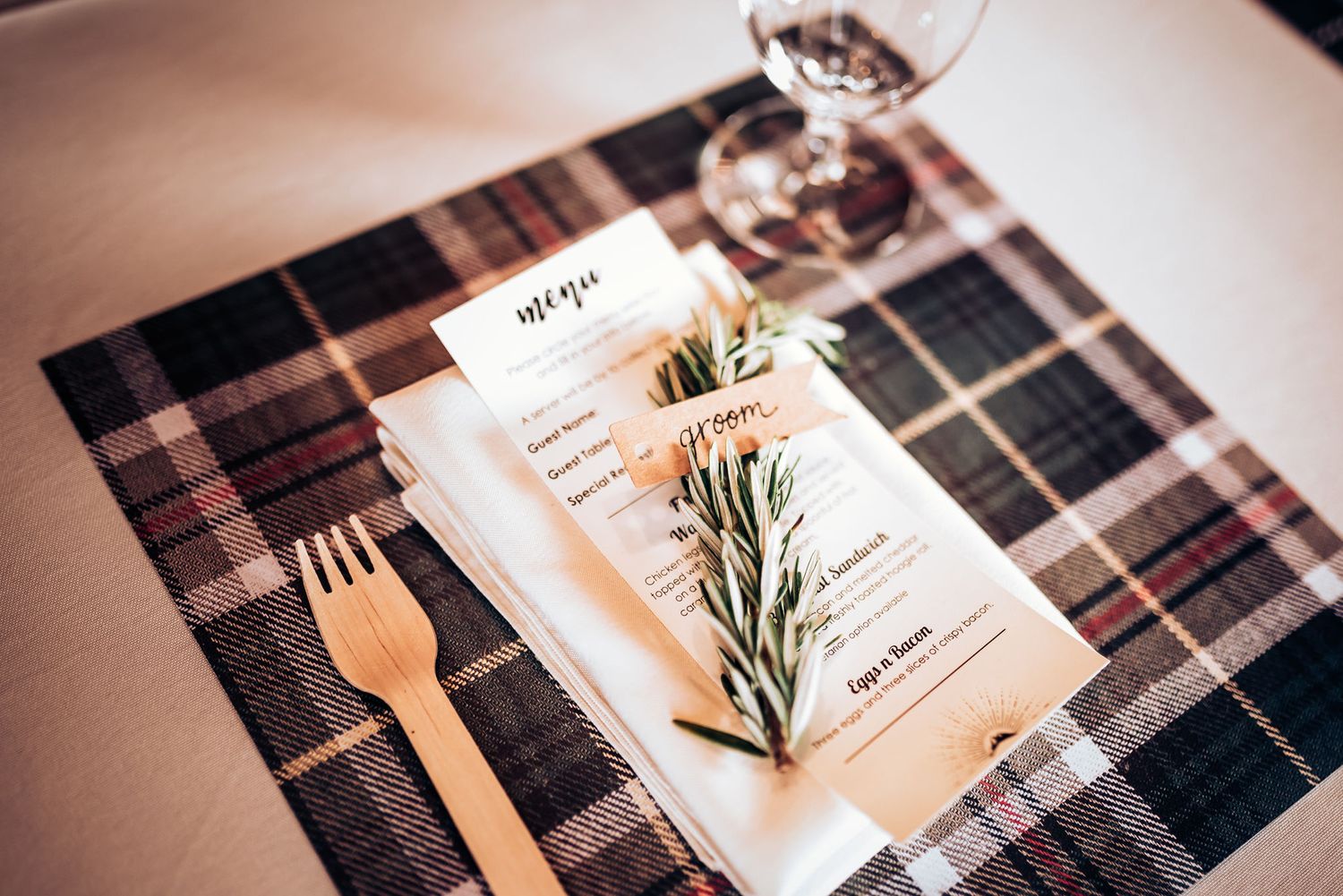 Wedding place setting with menu on plaid tablecloth decorated with rosemary sprig.