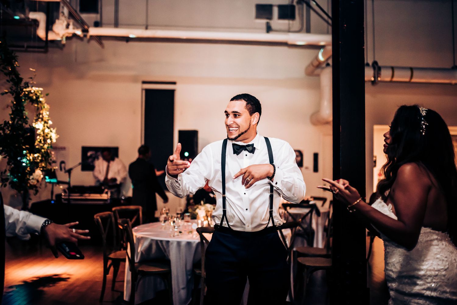 A person in suspenders and bow tie gestures enthusiastically during a wedding celebration.