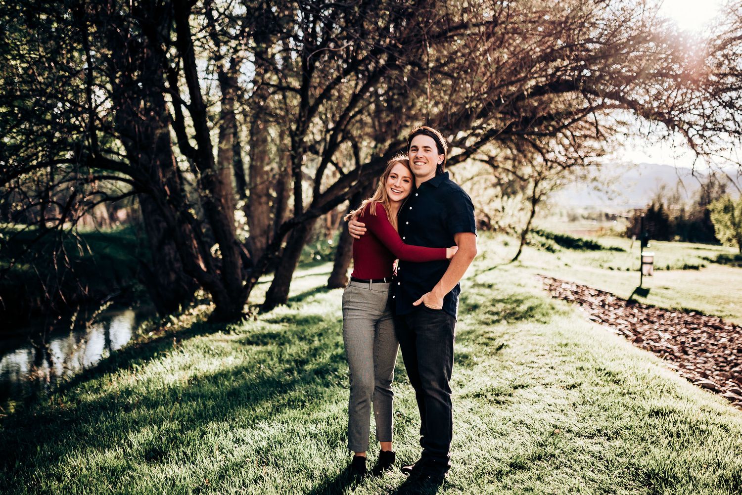 A couple stands together on a grassy path beside trees on a sunny afternoon.