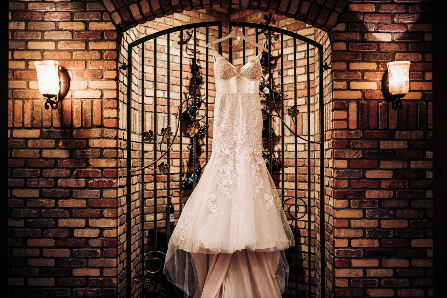 An elegant white wedding dress hanging in a brick archway with decorative iron gates.