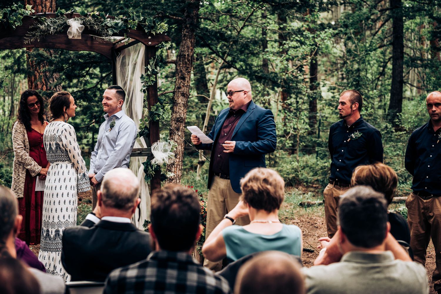 An outdoor wedding ceremony in a wooded setting with guests seated on chairs watching the proceedings.
