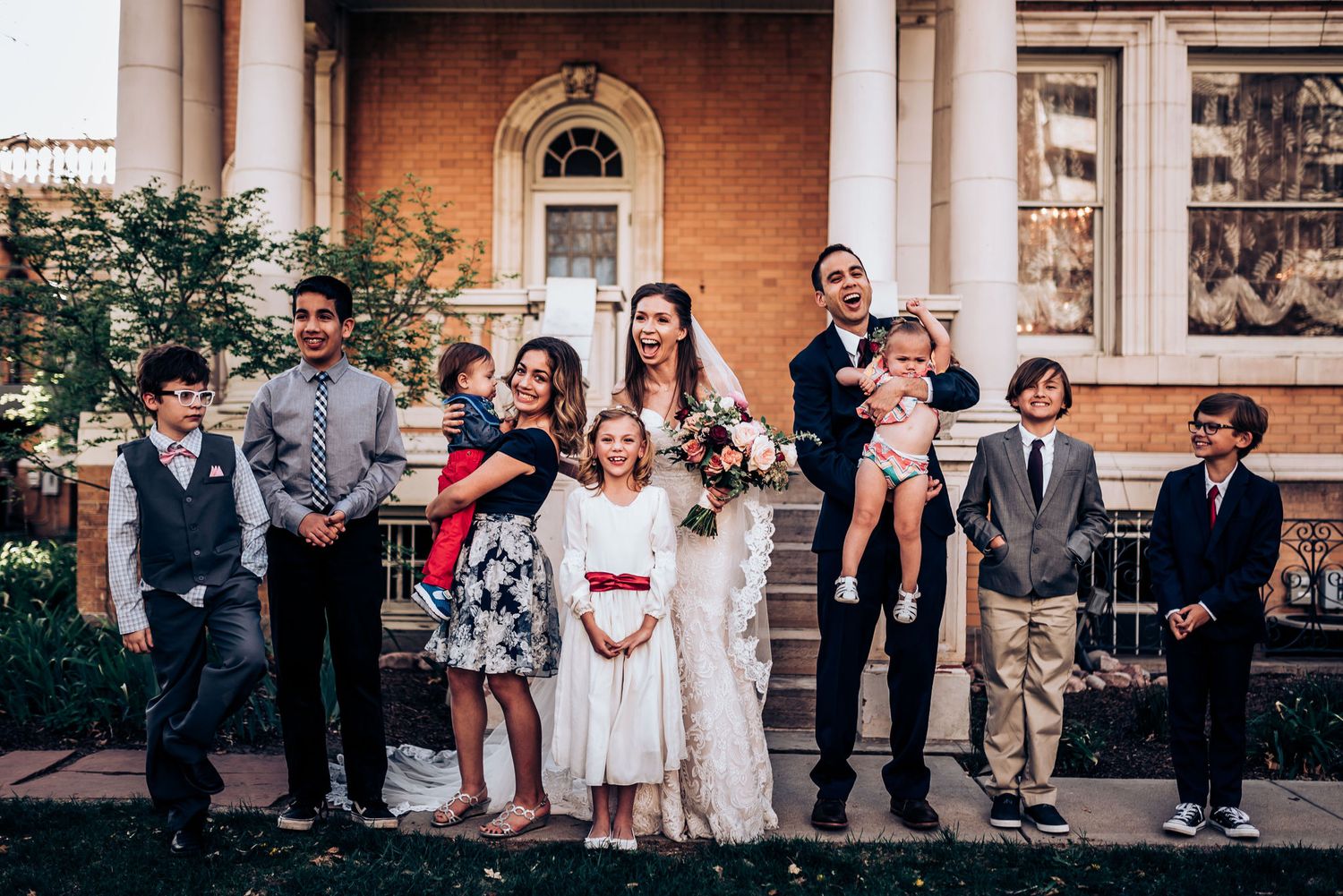 Large wedding group photo with children and adults outside historic brick venue.