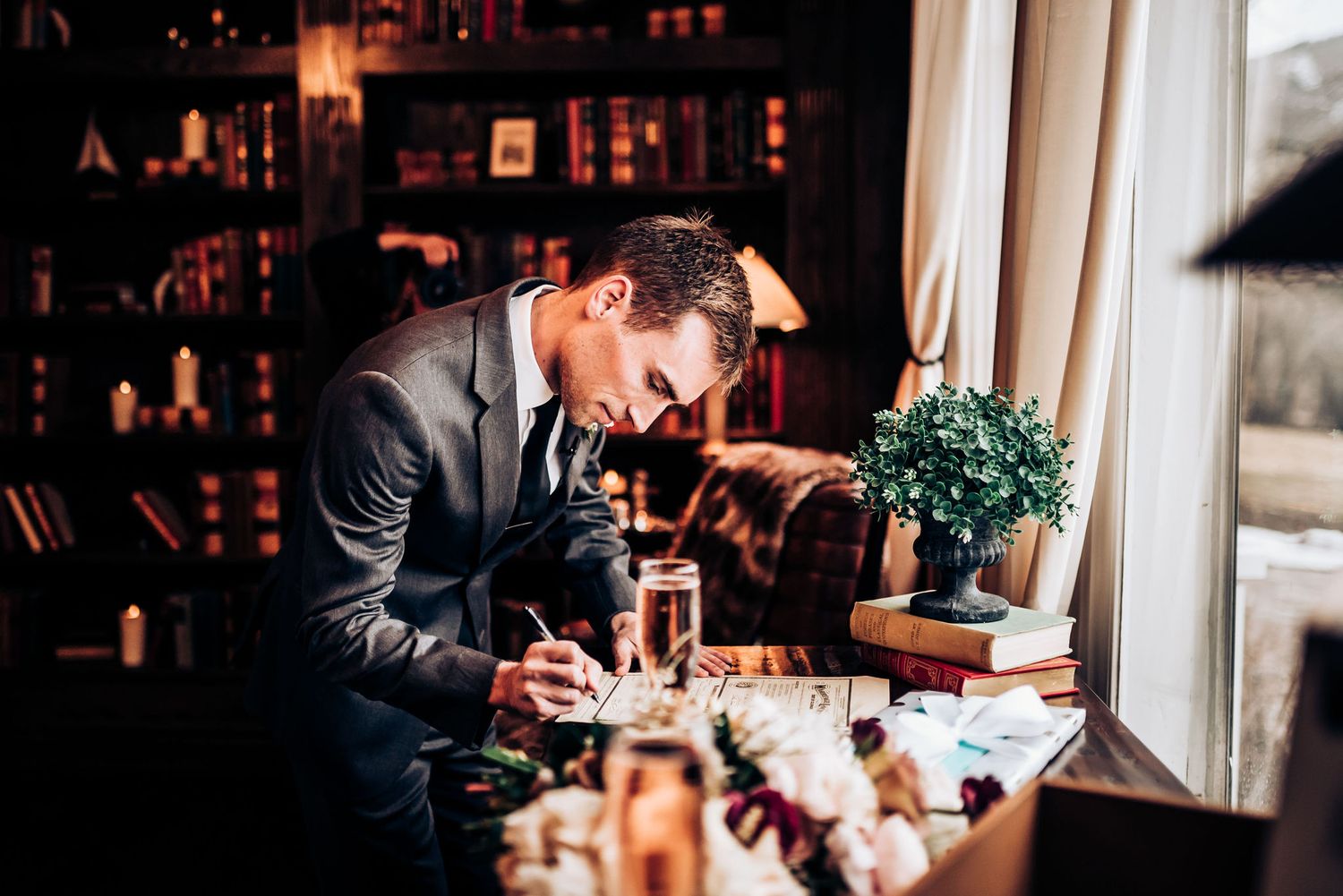 Formally dressed person writing at a window desk in a room with bookshelves and warm lighting.