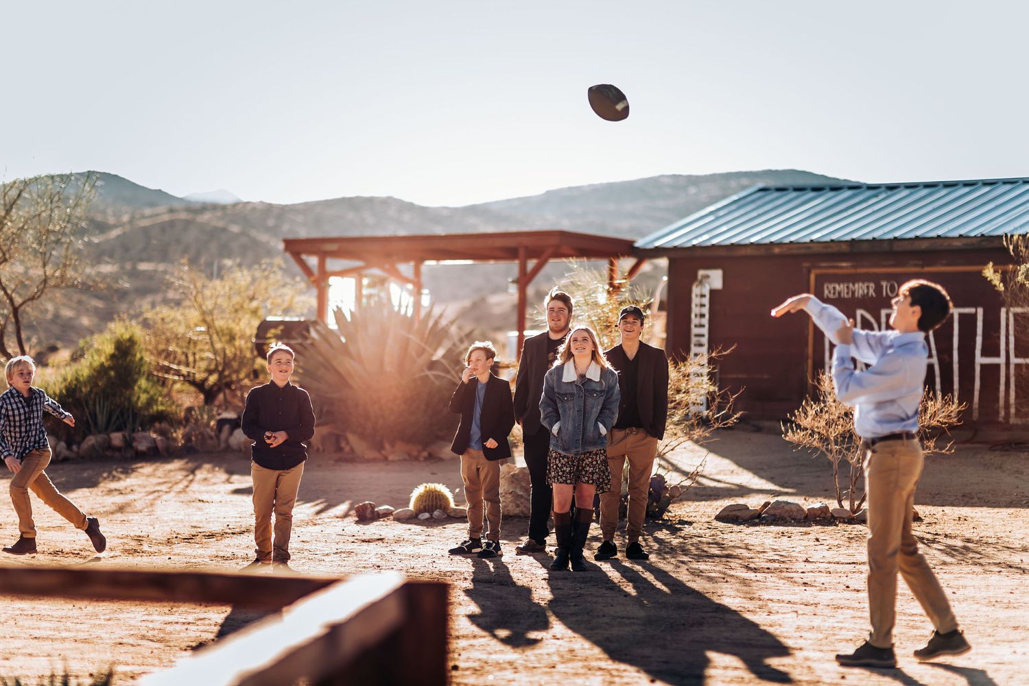 People playing football outside a desert cabin during sunset with mountains in background.