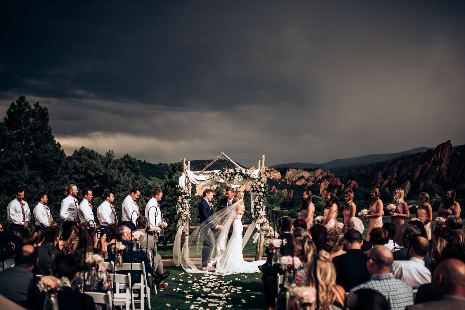 Wedding ceremony at dusk with guests seated on white chairs and mountain landscape in background.