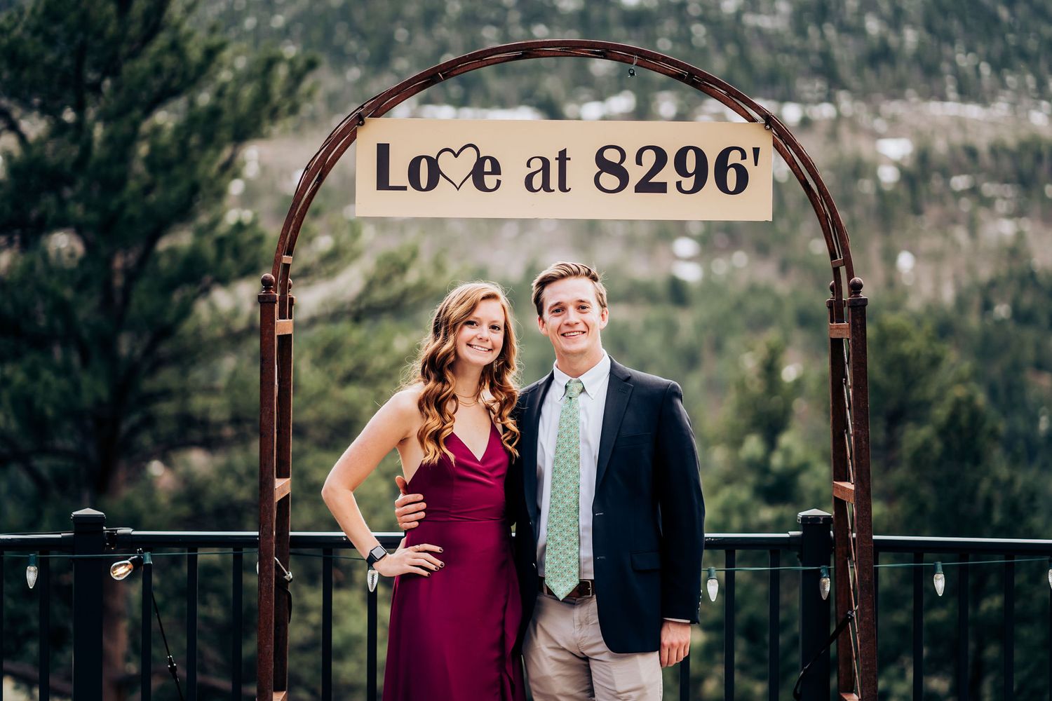 A couple poses under an arch with 'Love at 8296' sign against a mountainous backdrop.