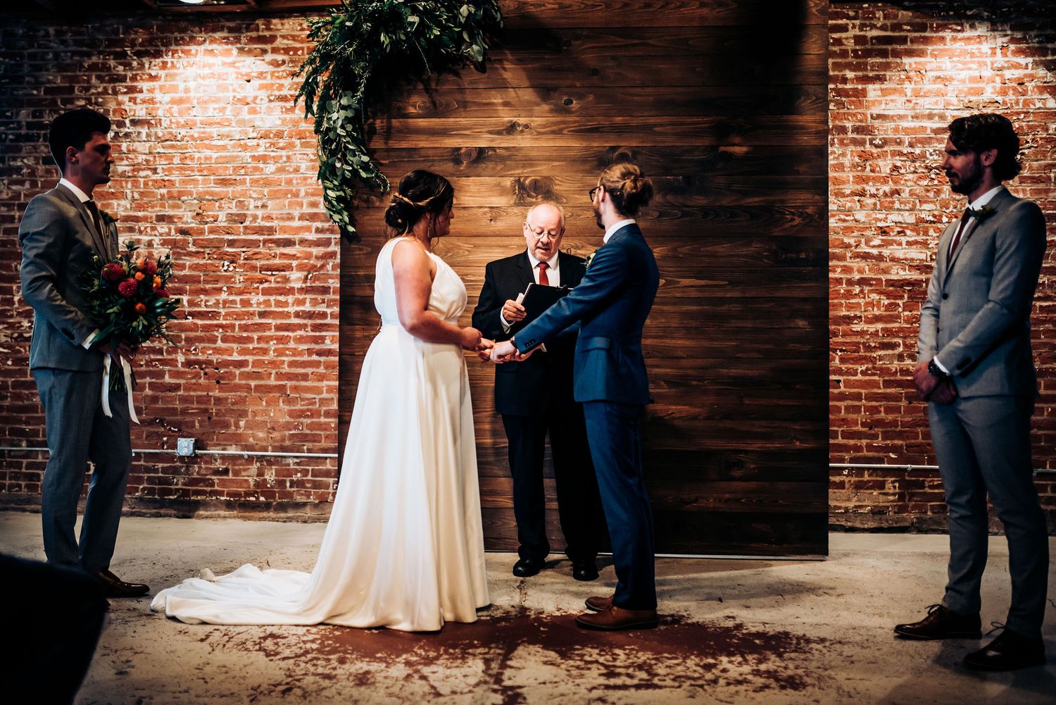 Wedding ceremony takes place in front of wooden accent wall with greenery and exposed brick details.