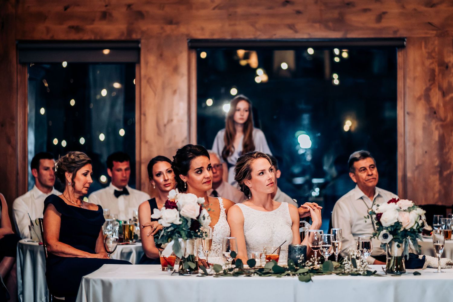 Wedding party seated at decorated head table during evening reception with twinkling lights.