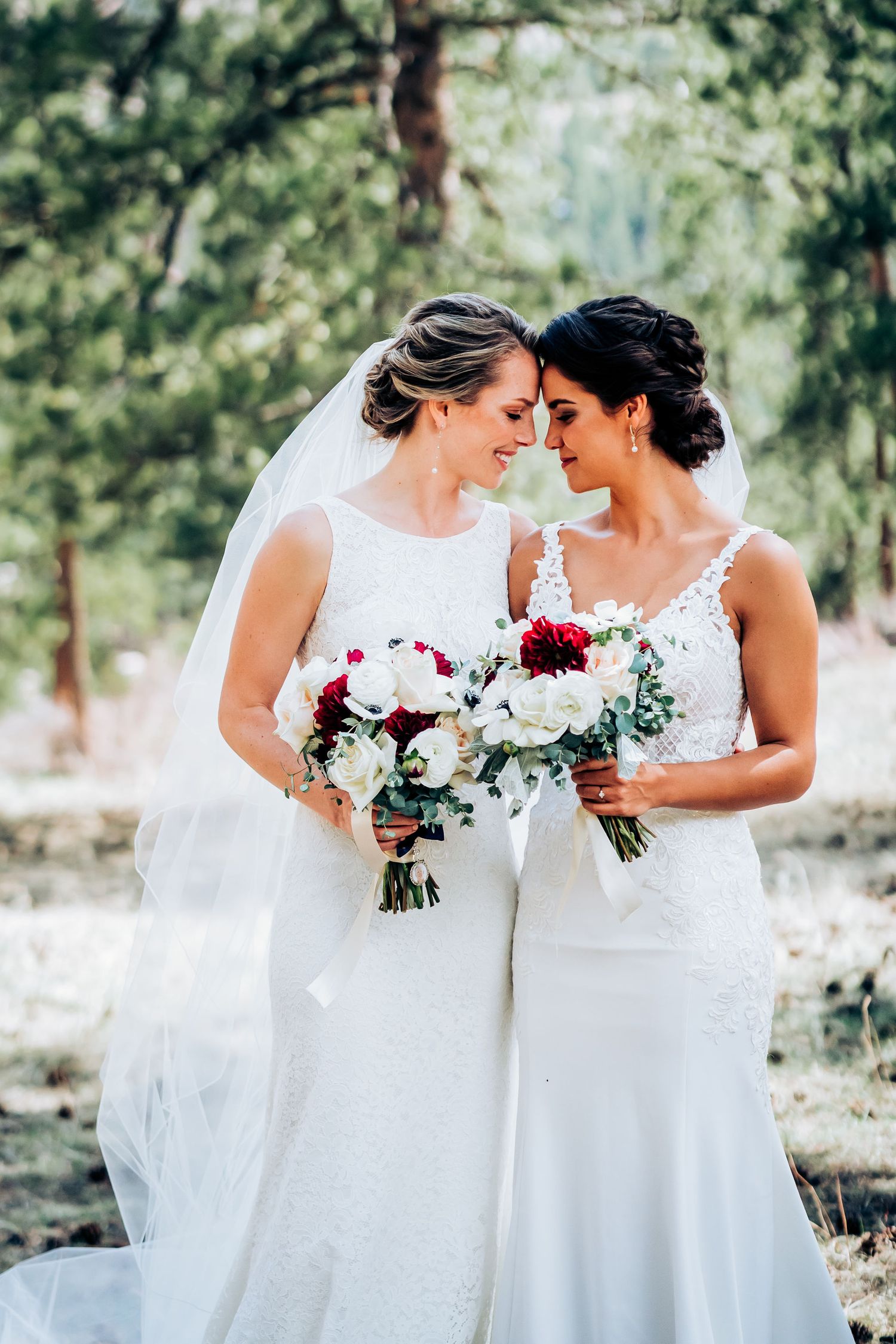 Two brides share an intimate moment in the forest while holding elegant white and red bouquets.
