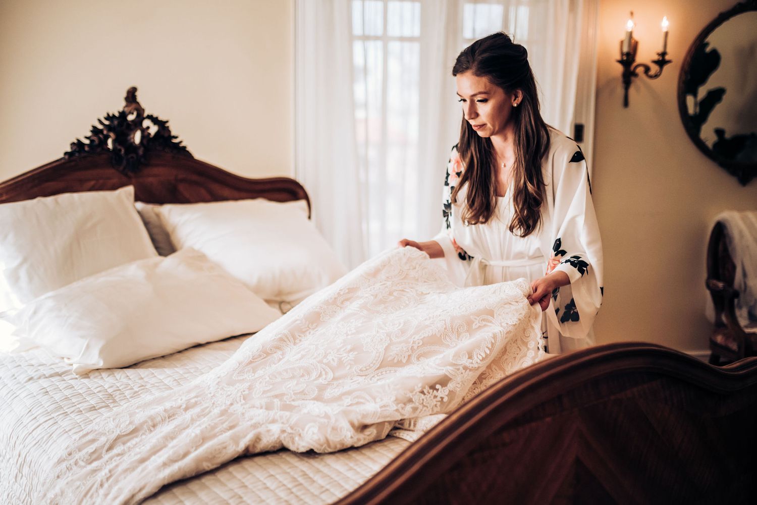 A delicate lace wedding dress displayed on an ornate wooden bed with soft natural lighting.