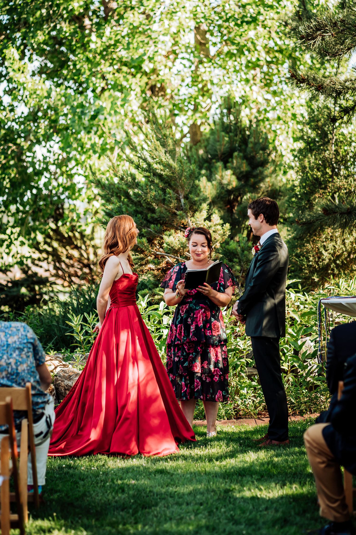 A wedding ceremony takes place in a lush garden setting with attendees in formal attire.