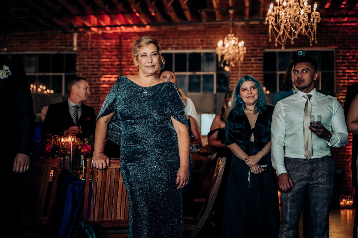 Wedding guests in formal attire gather in an elegant brick venue with crystal chandeliers.