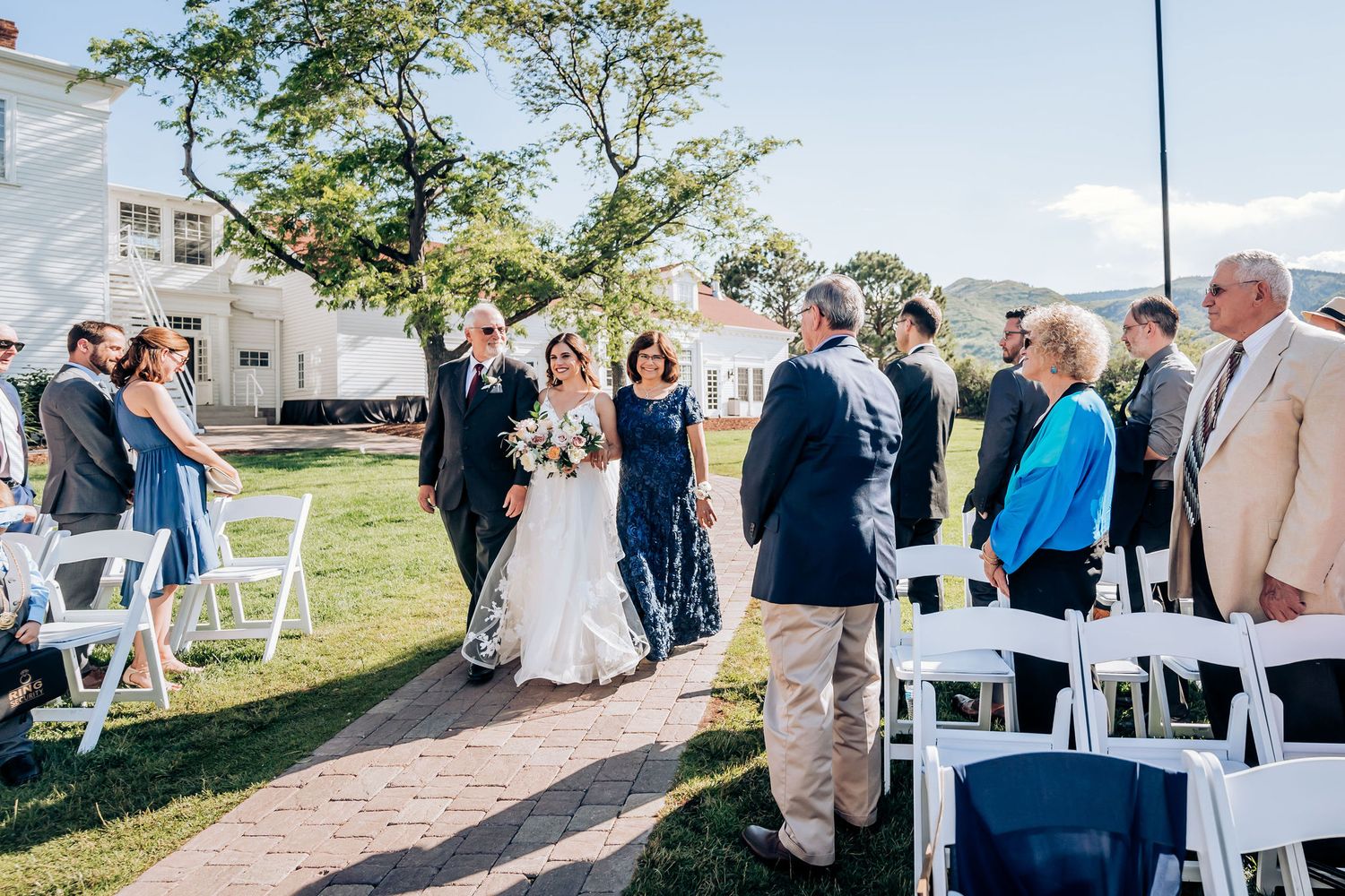 Wedding ceremony processional on stone pathway with white chairs and colonial style building.
