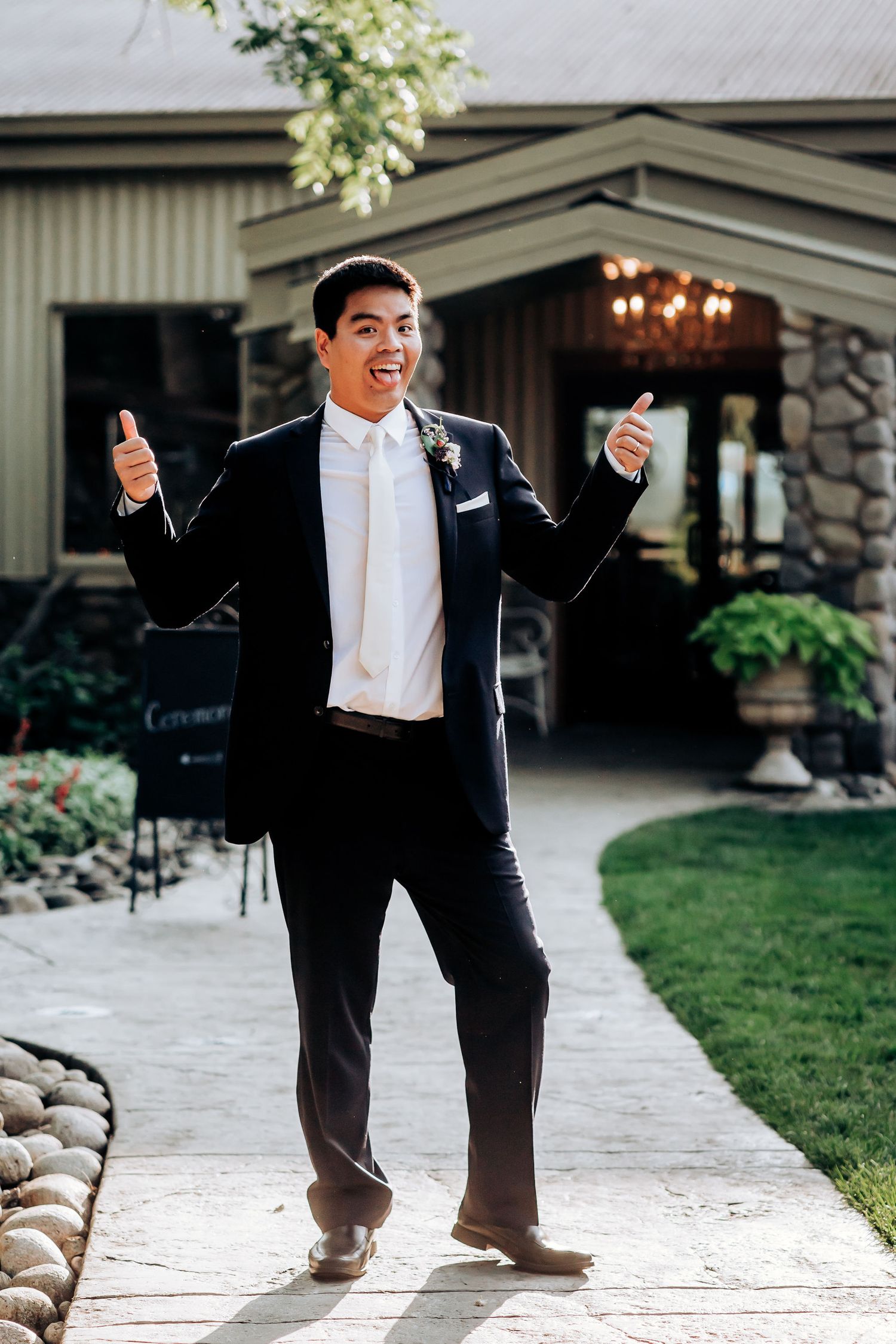 Excited groom in black tuxedo giving thumbs up outside venue entrance.