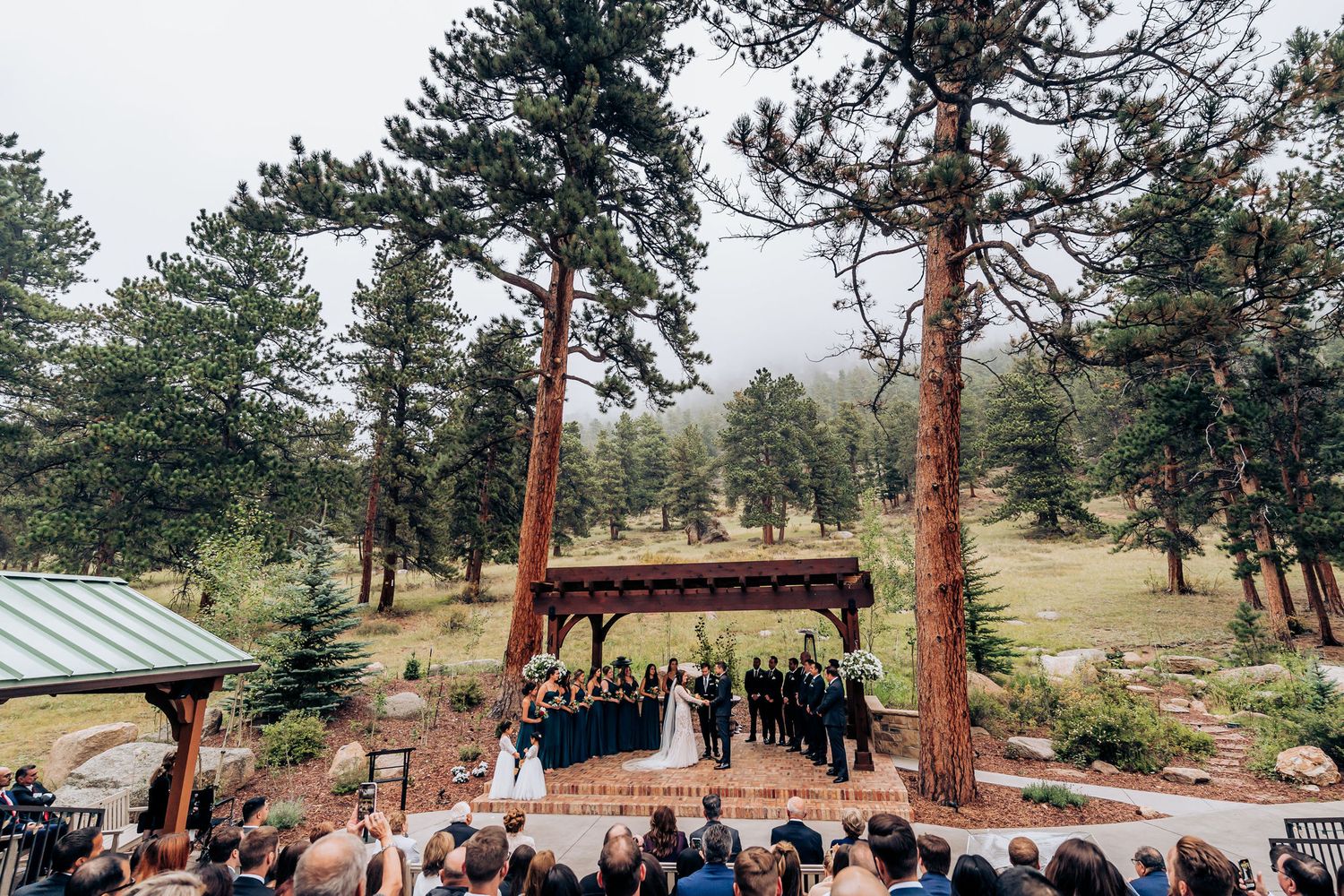 An outdoor wedding venue features a wooden gazebo nestled among tall pine trees in a mountain setting.