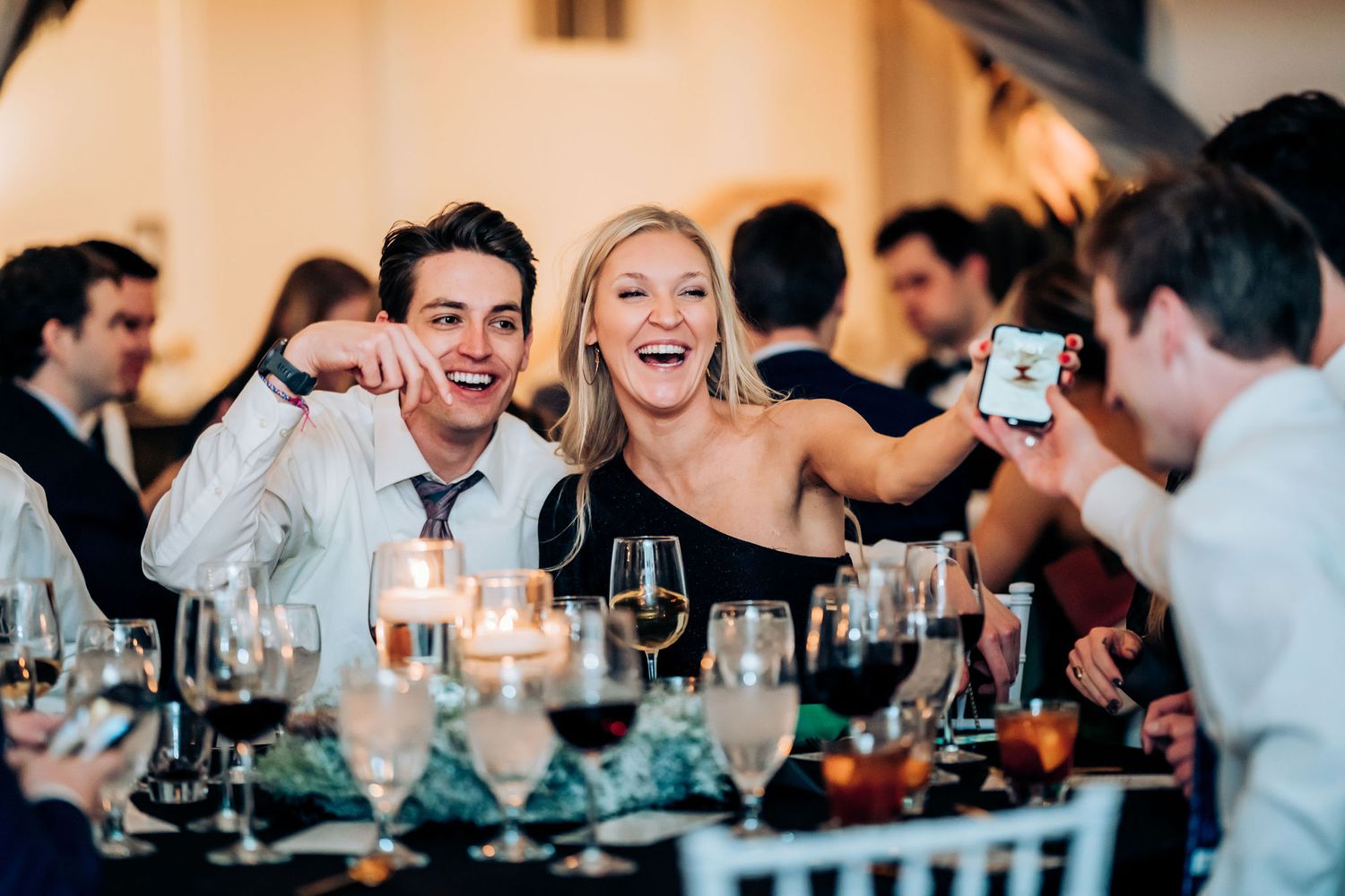 Wedding reception guests share laughter and joy around an elegantly set dinner table with wine glasses and candlelight.
