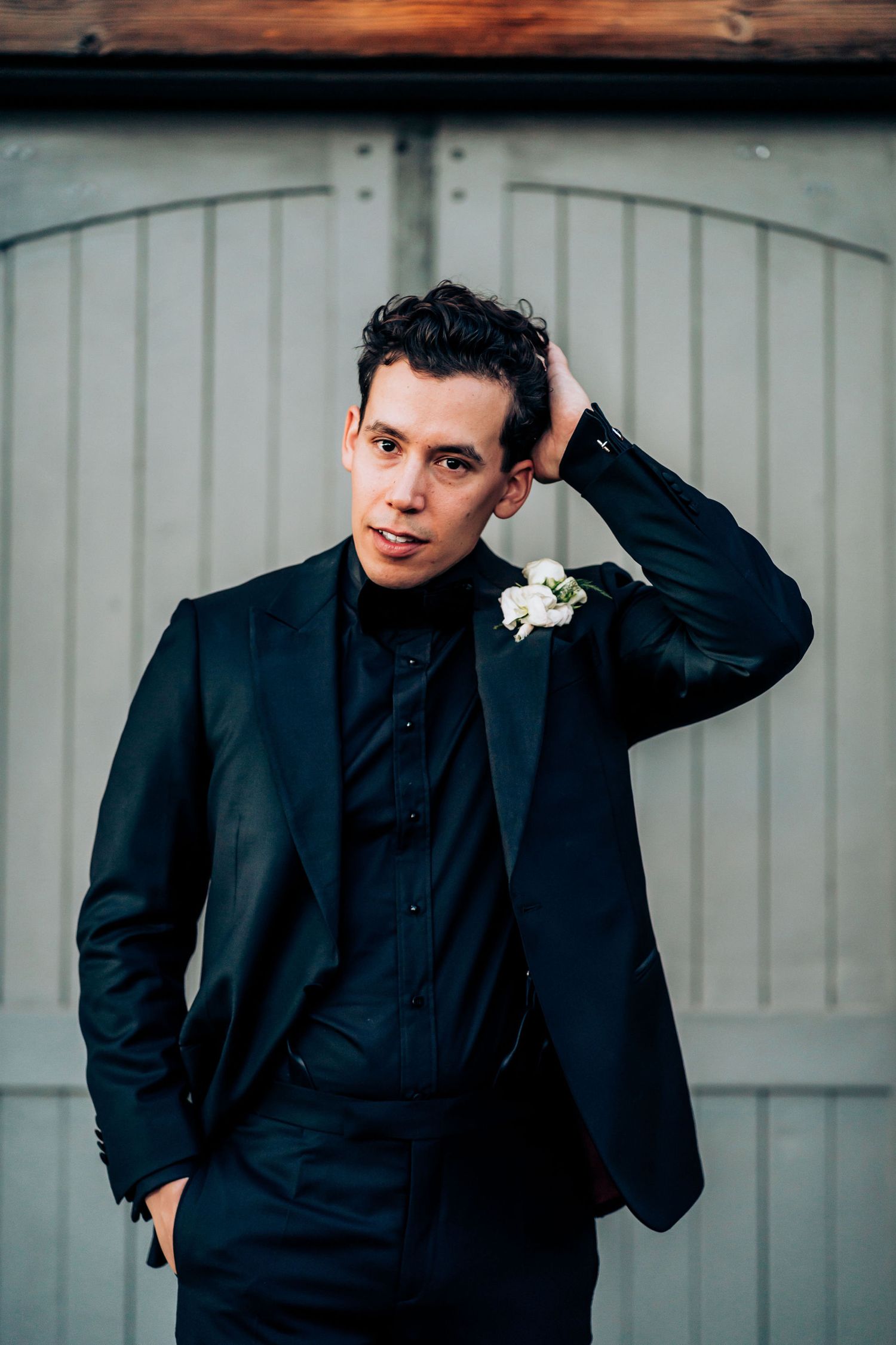 Groom poses in a sleek navy suit with a white boutonniere against a light-colored barn door background.
