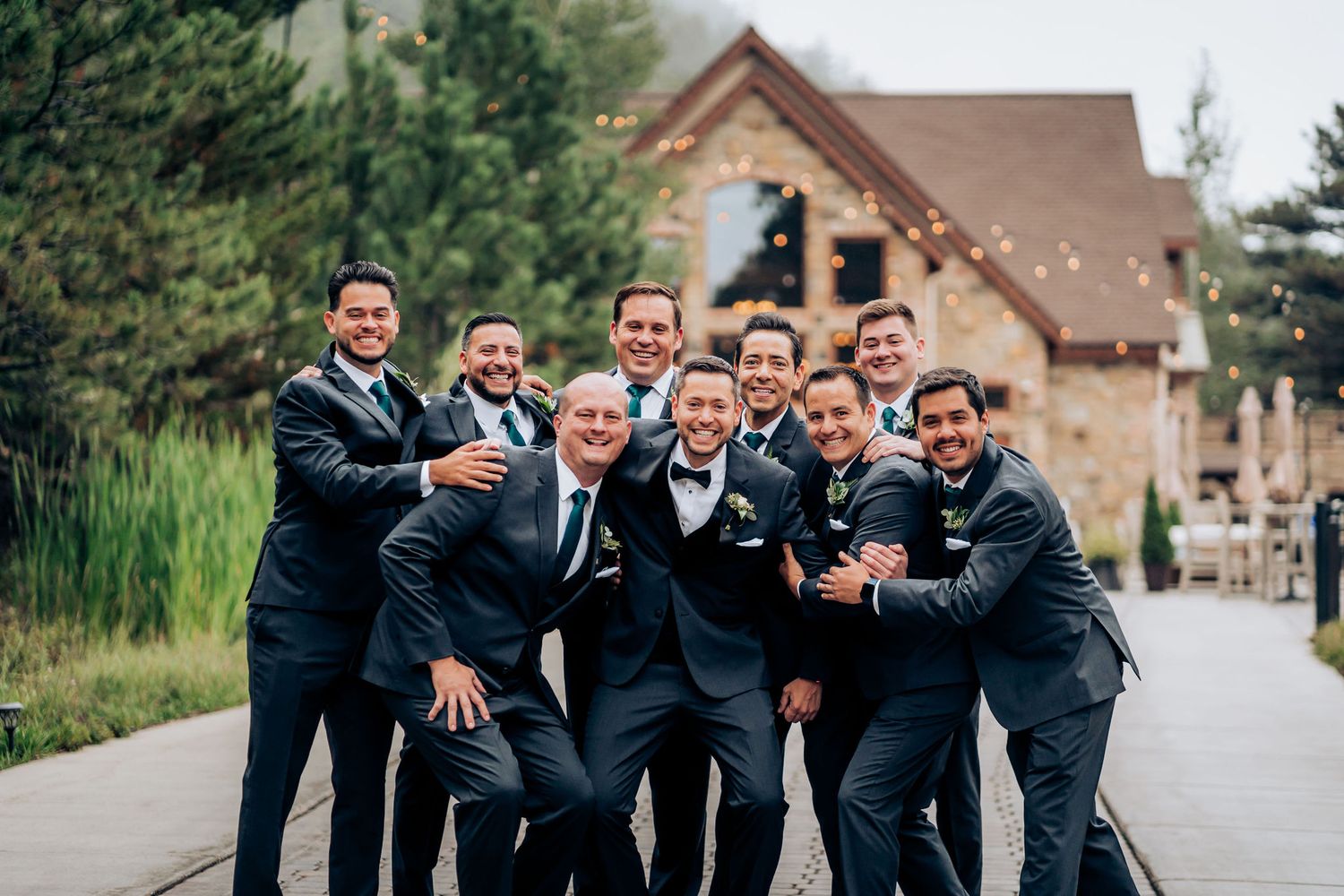 Groomsmen in navy suits pose playfully outside a stone and wood venue.