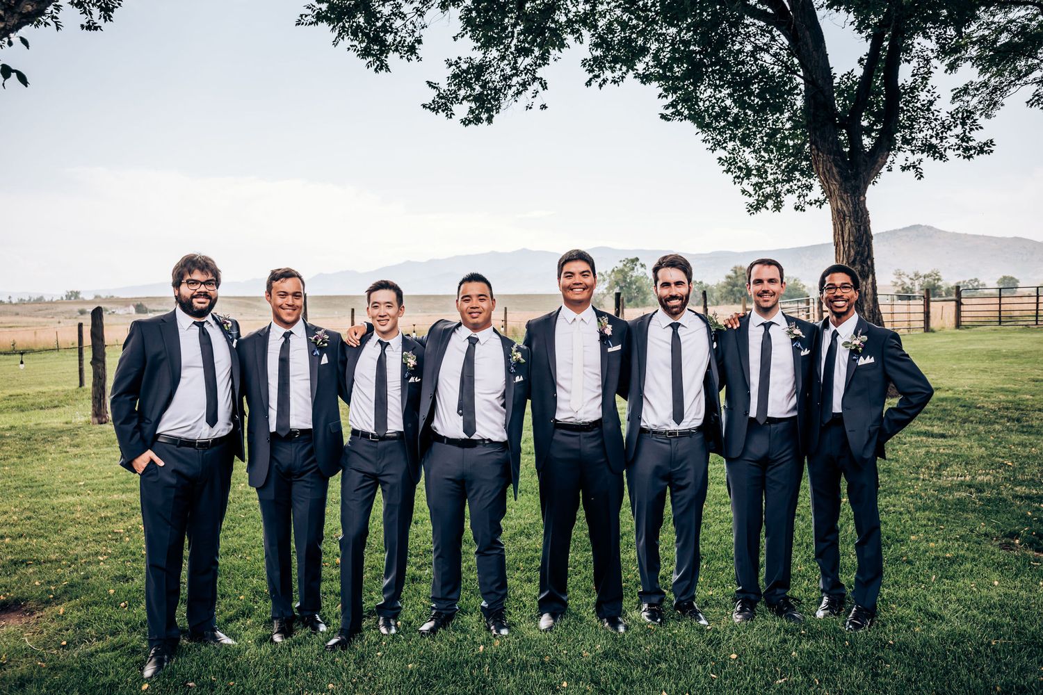 Groomsmen in navy suits and black ties pose together on grass with mountains in background.