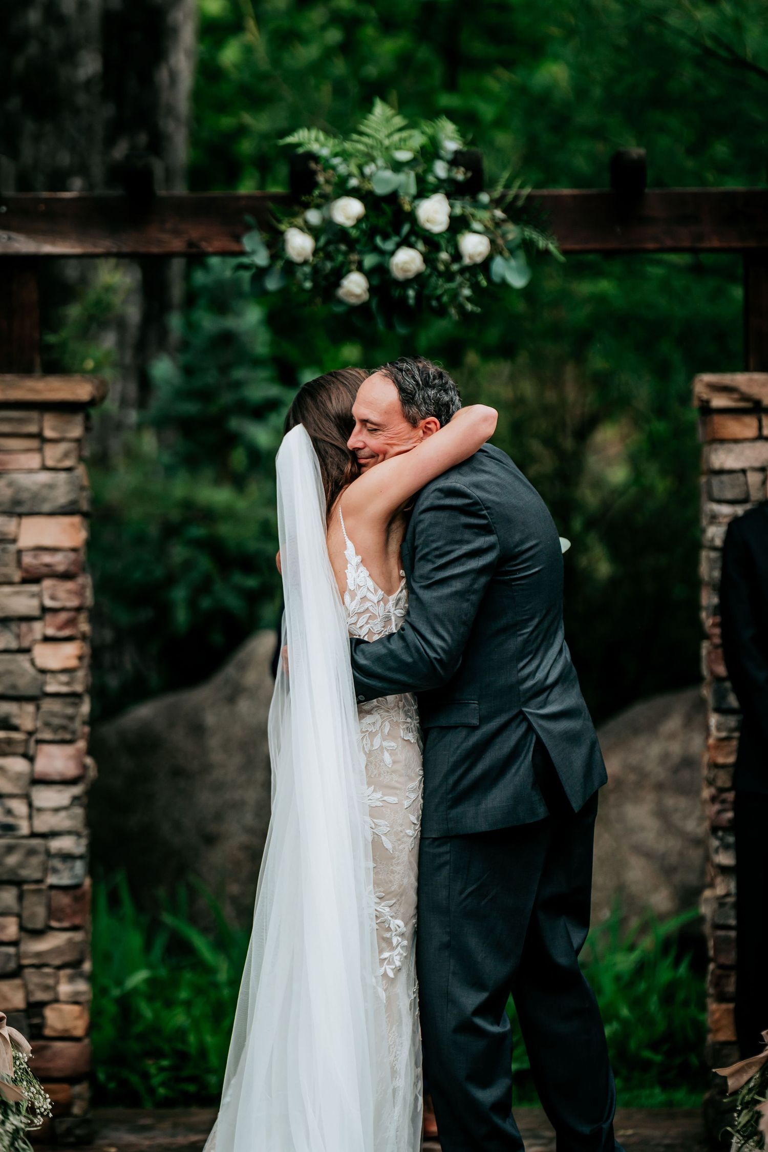Newlyweds share a romantic kiss under a floral arch with stone pillars.