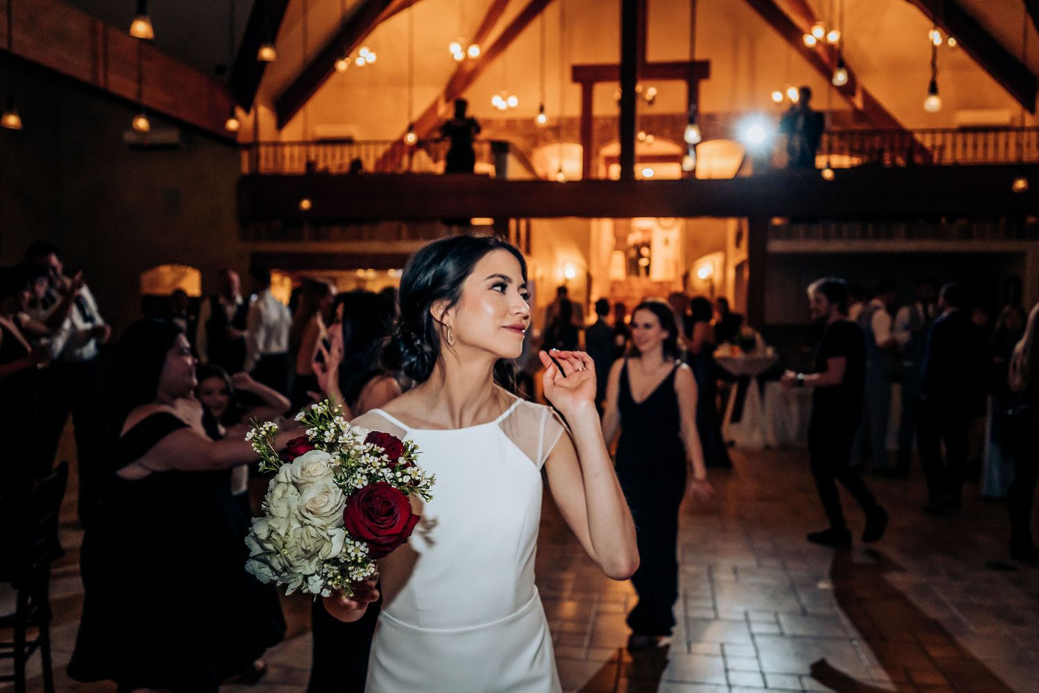 Person holding a bouquet sings during a wedding reception in a rustic venue with vaulted ceilings.