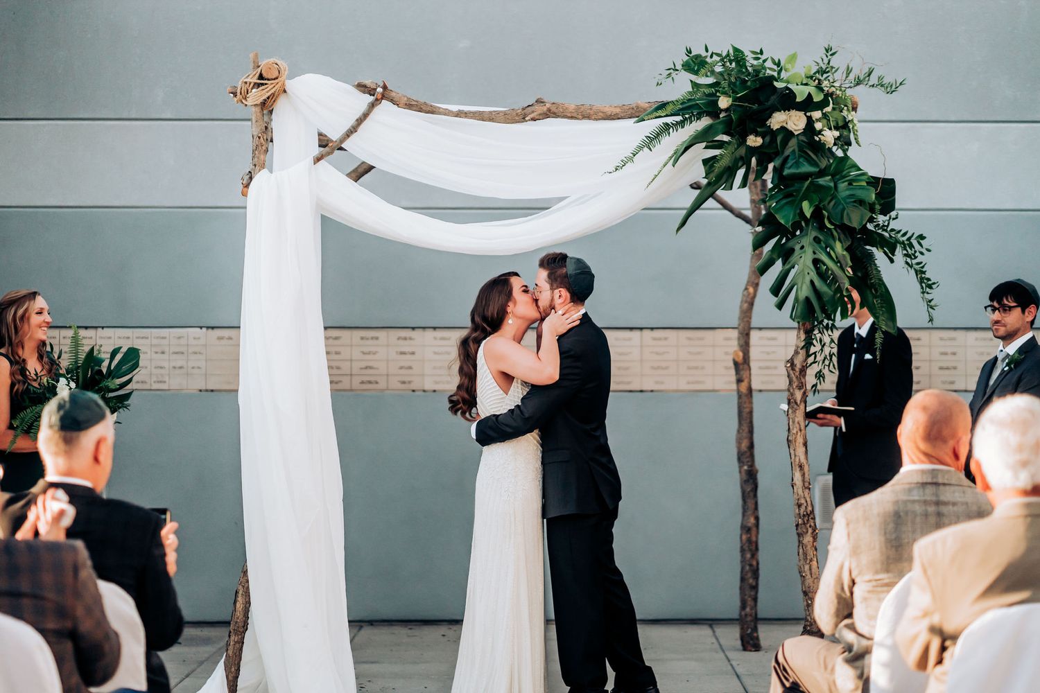 Wedding ceremony moment under a white draped arch with floral decorations and guests watching.
