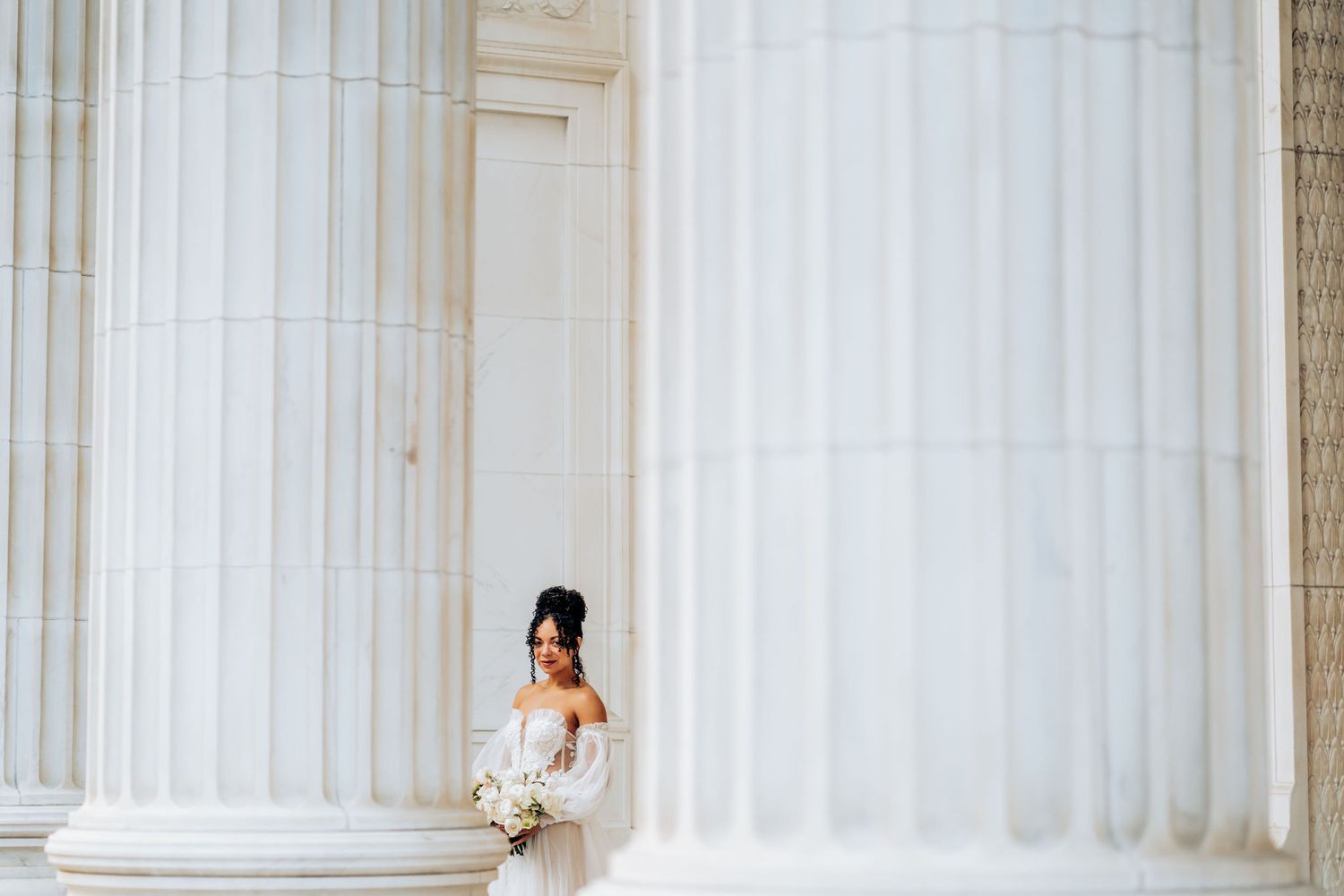 Bride in white dress holding bouquet poses against classical white stone columns.