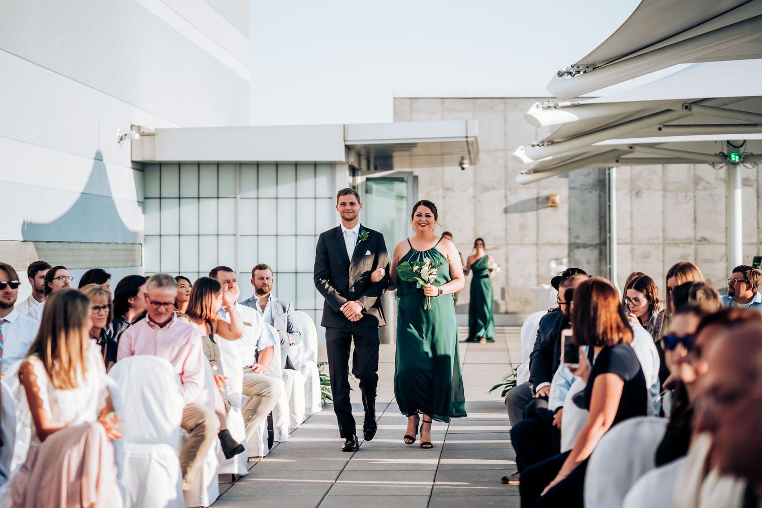Attendants in dark suits and a green dress walk down an outdoor wedding aisle while guests watch from white chairs.