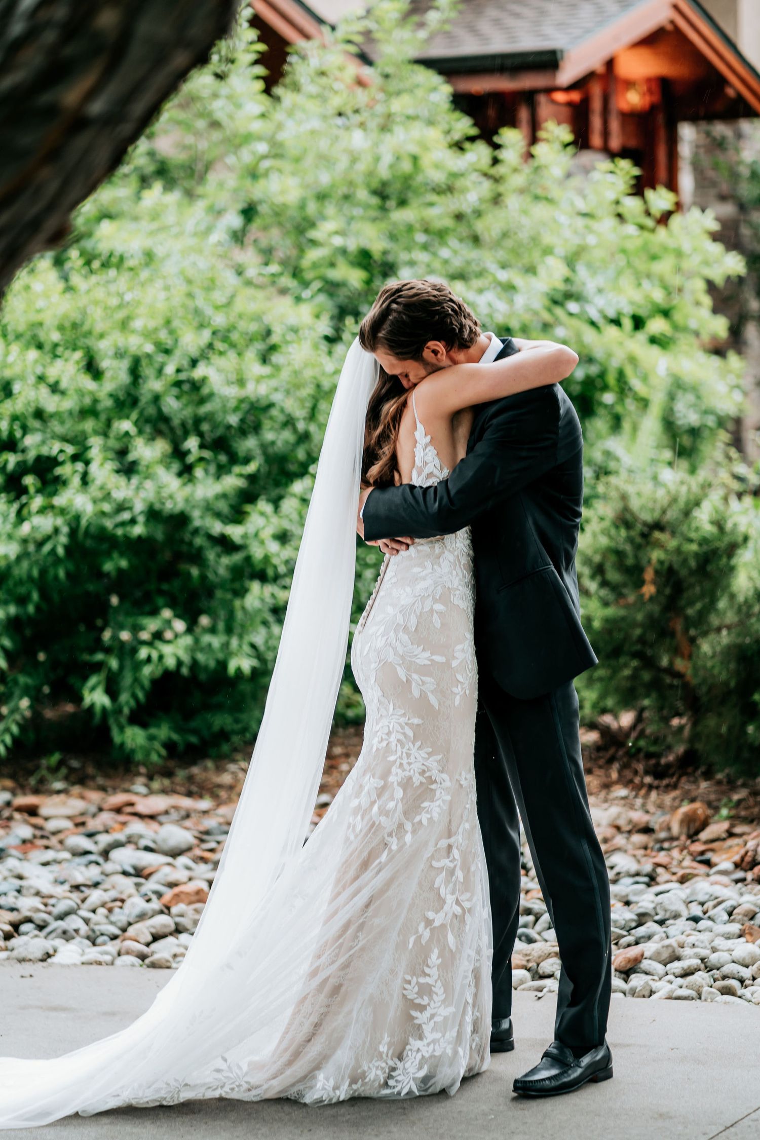 Elegant wedding embrace under covered entrance with flowing lace dress and long veil.