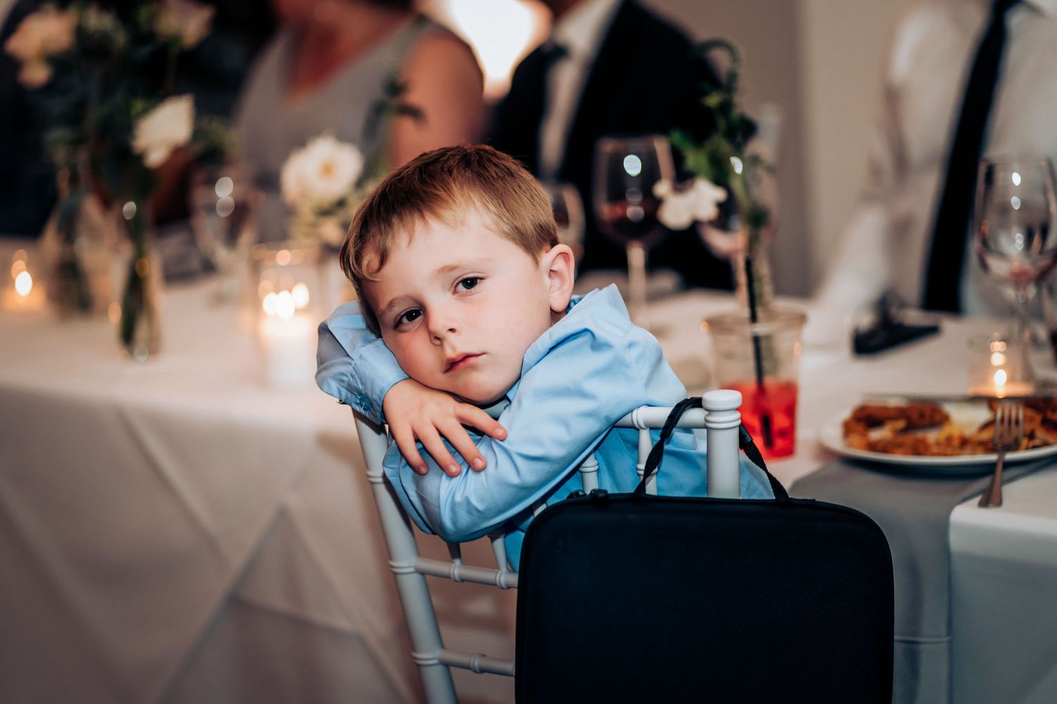A young boy in light blue attire rests his head on a chair during the wedding reception.