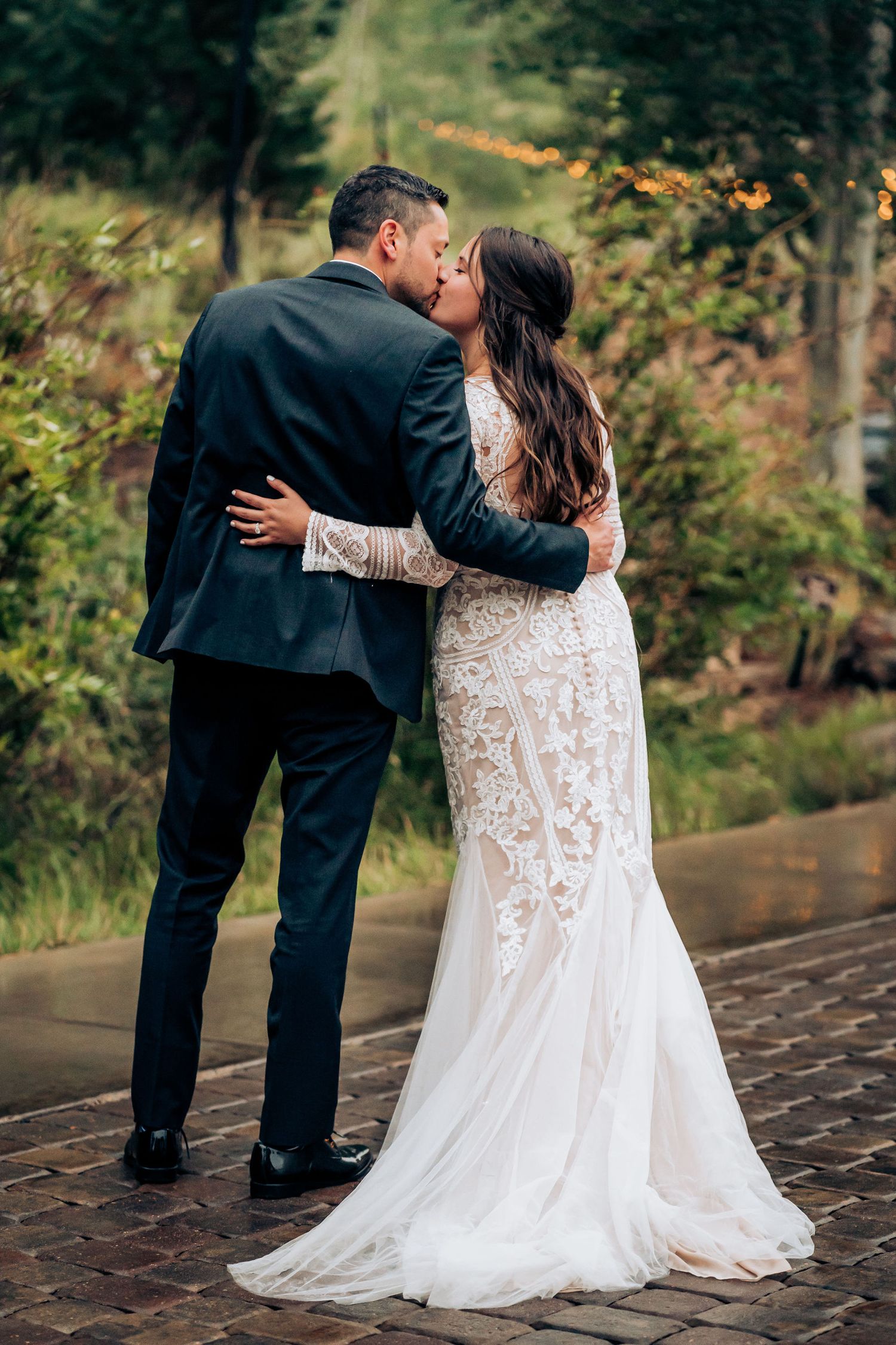 Romantic wedding couple embracing on a brick pathway with string lights and greenery.