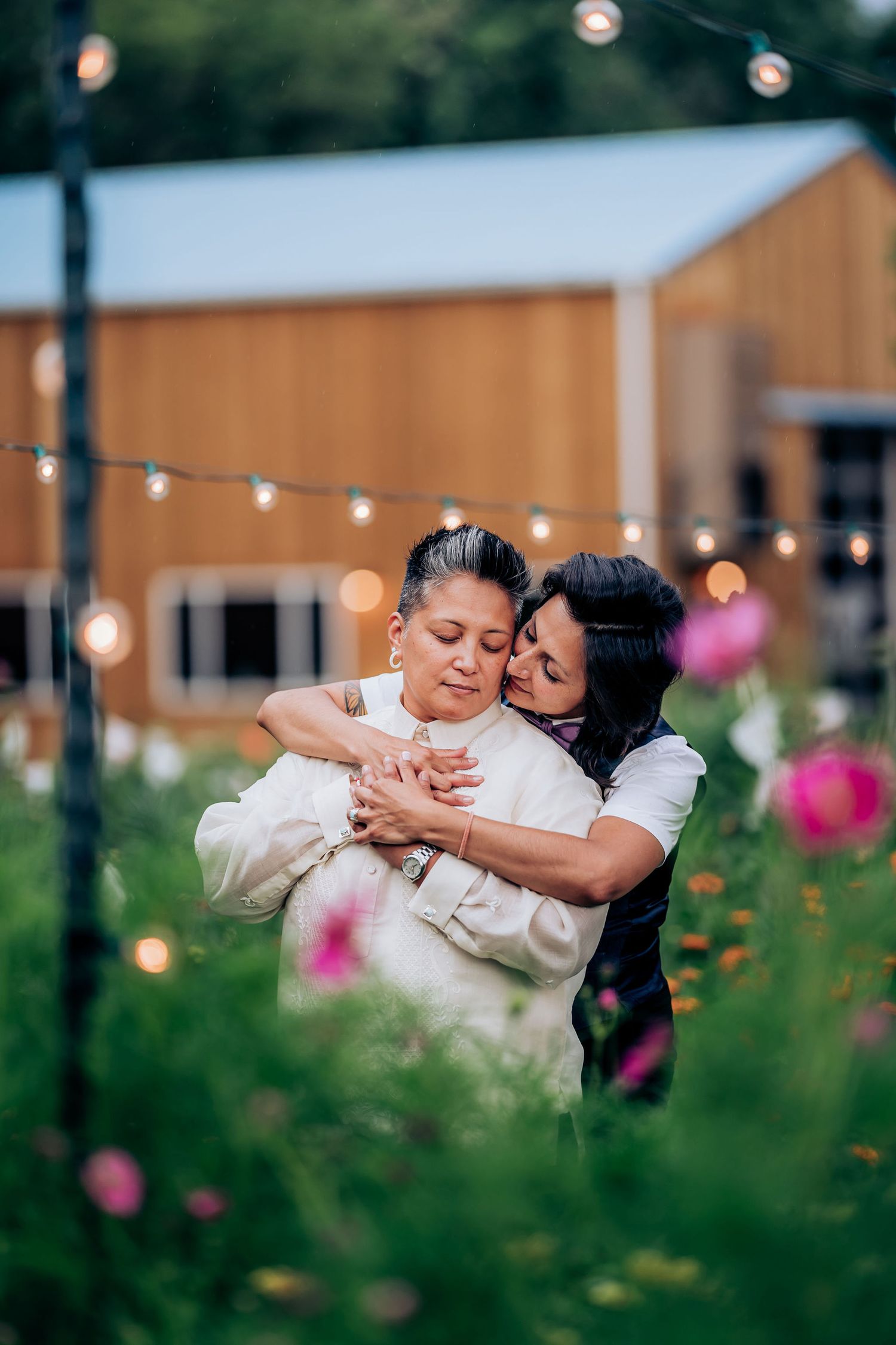 Two people sharing a romantic moment among pink flowers with a rustic barn in the background.