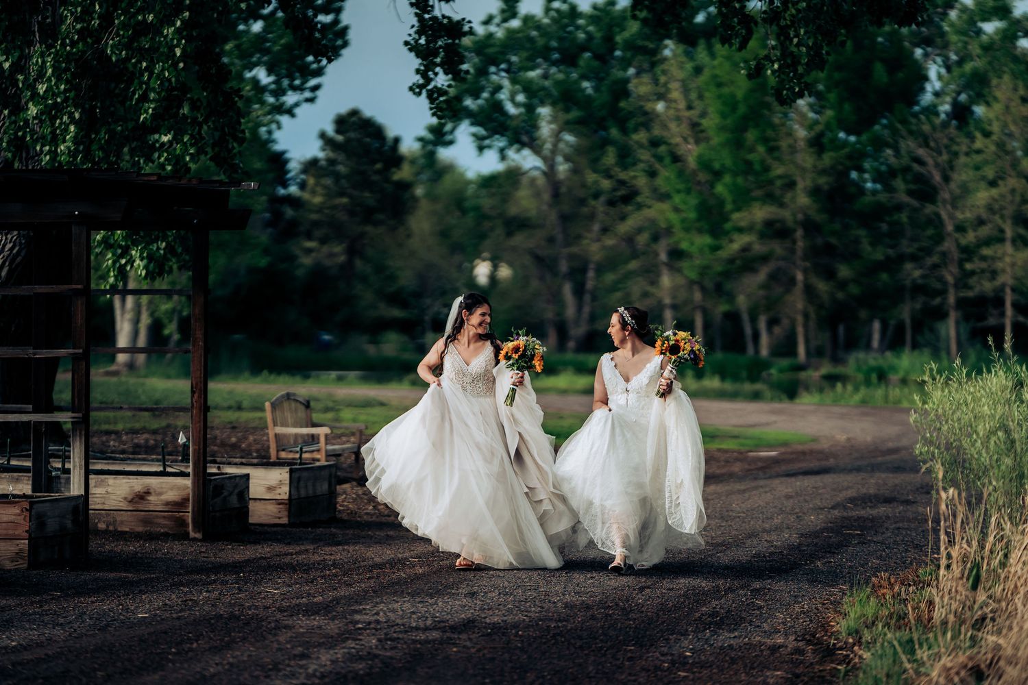 Two people in white wedding gowns run joyfully down a dirt path surrounded by trees.