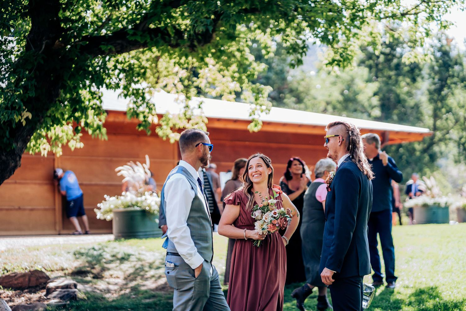 Outdoor wedding gathering with attendees in formal attire mingling under trees.