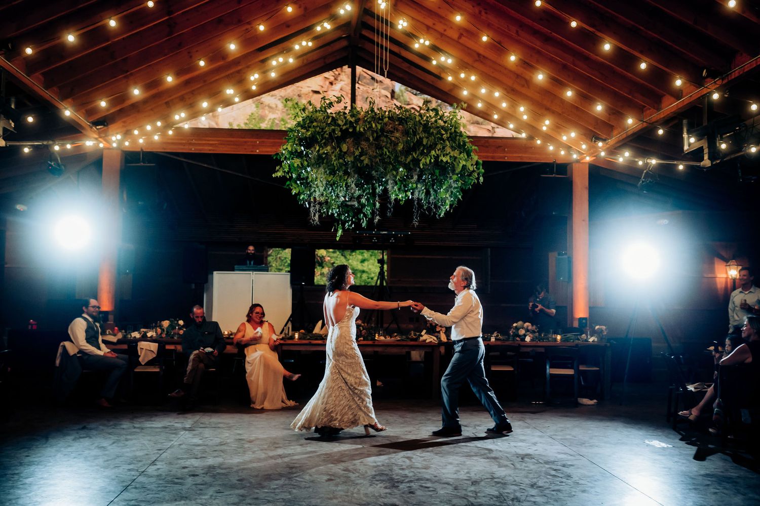 A couple dances under warm string lights in a rustic reception venue.