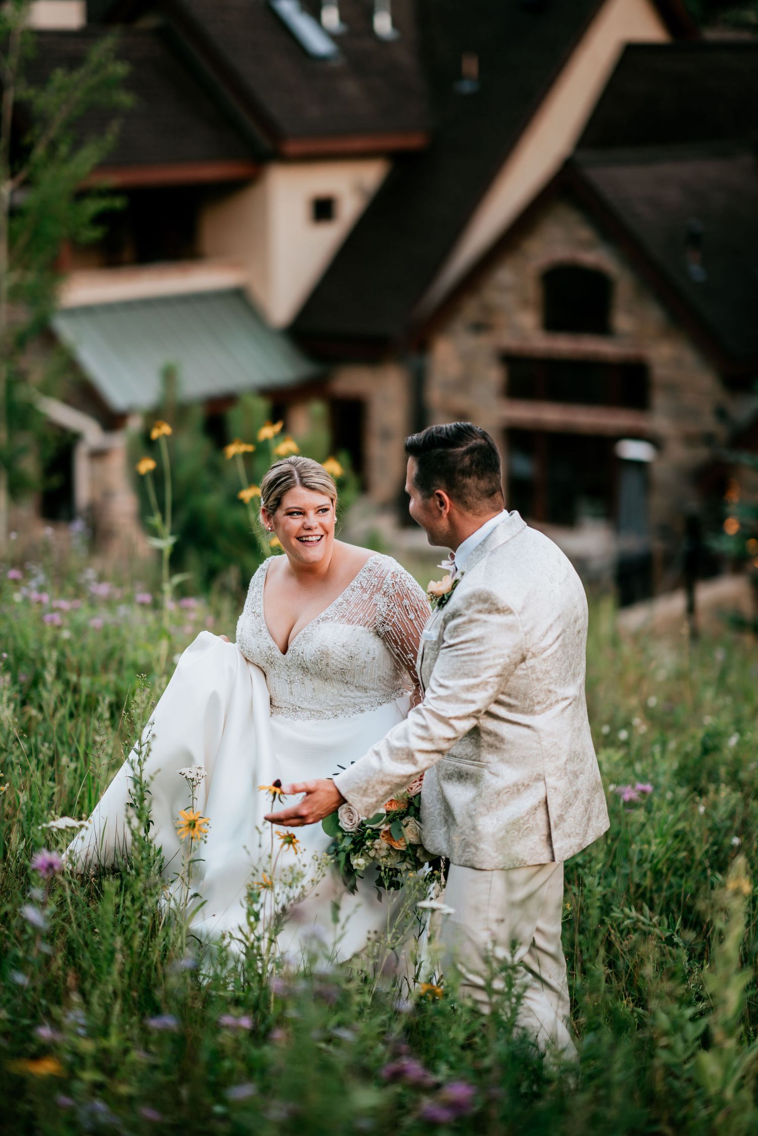 A couple in wedding attire shares a moment among wildflowers with a rustic house in the background.