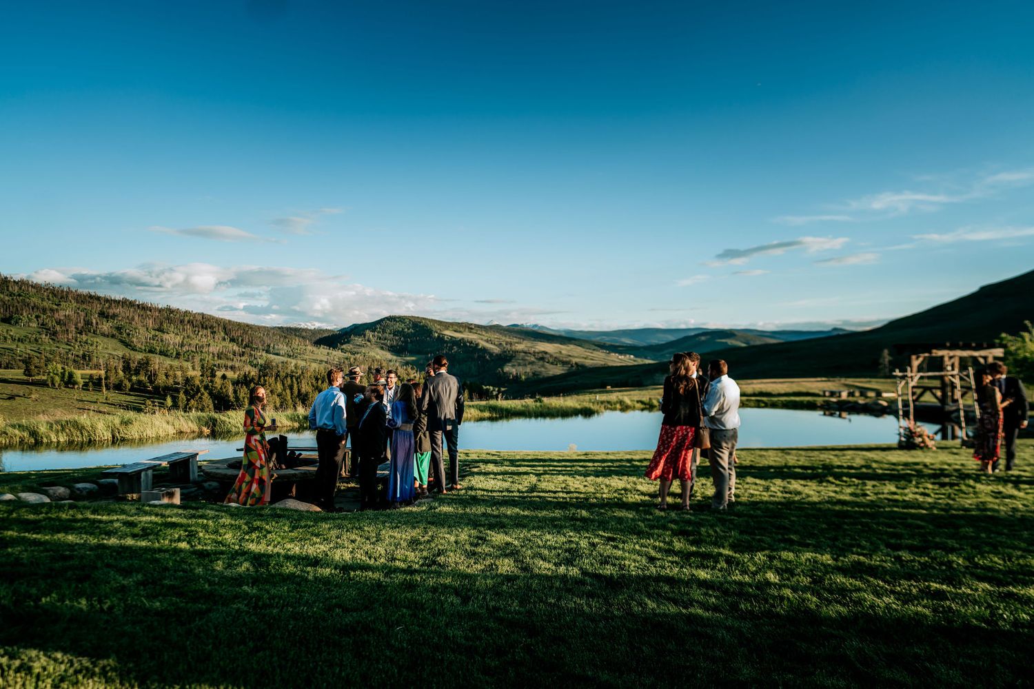 Wedding guests gather on scenic hillside overlooking mountain lake during outdoor ceremony.
