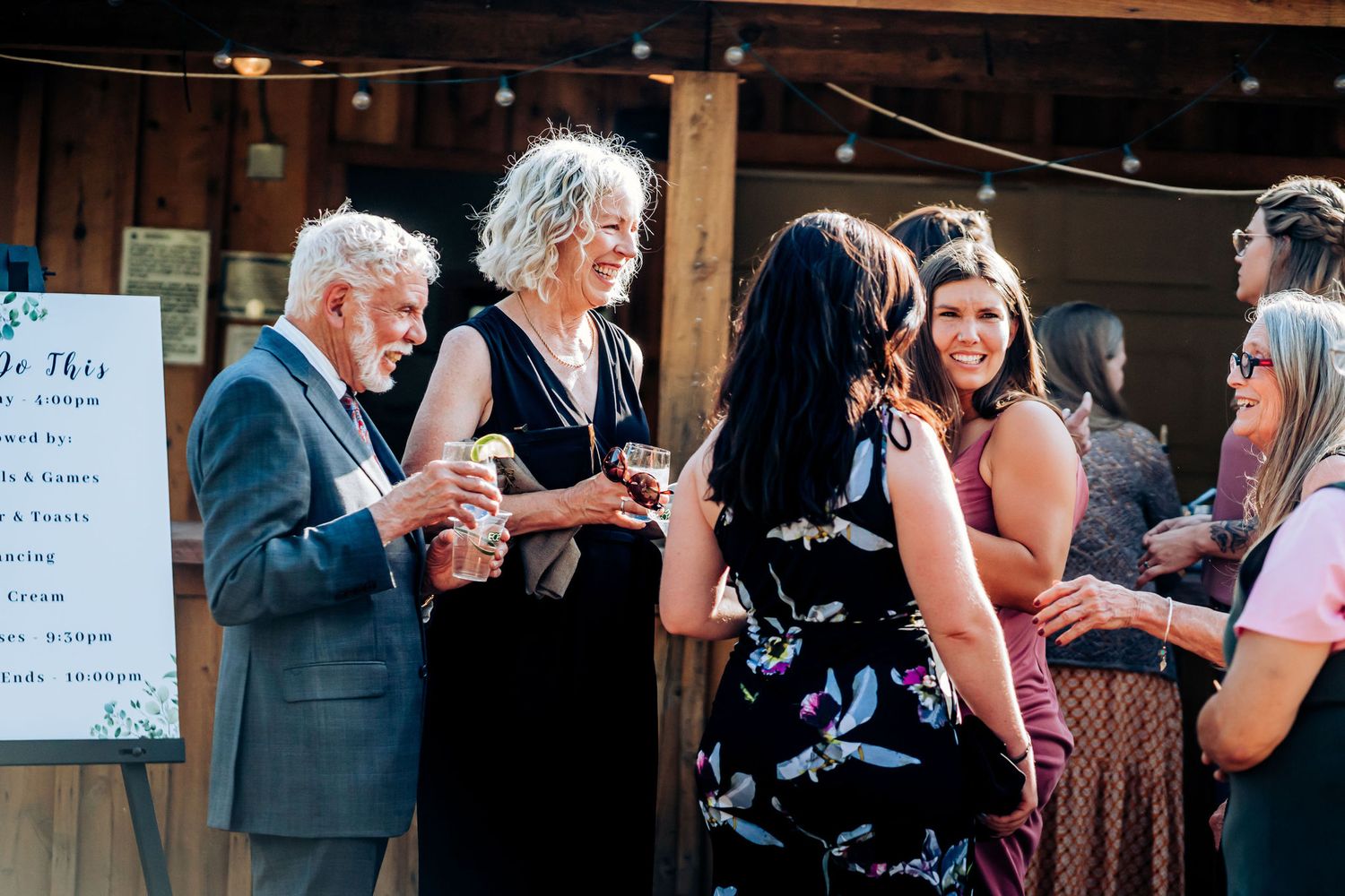Wedding guests mingling by event schedule sign board at rustic venue.