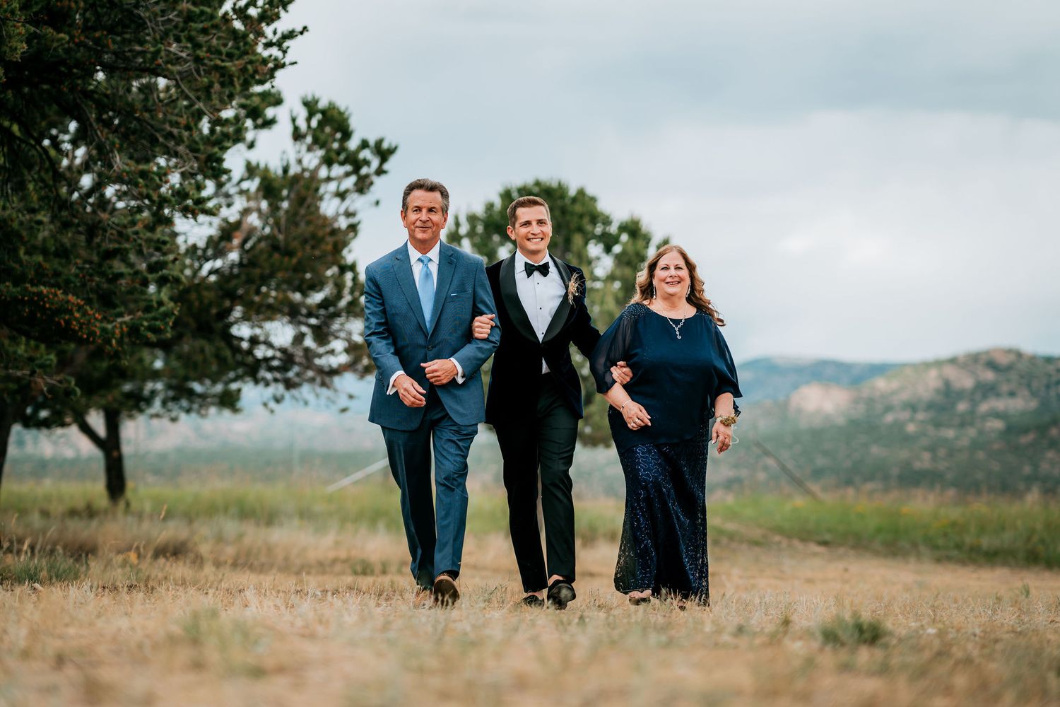 Three people walk arm-in-arm through a scenic outdoor setting during a formal event.